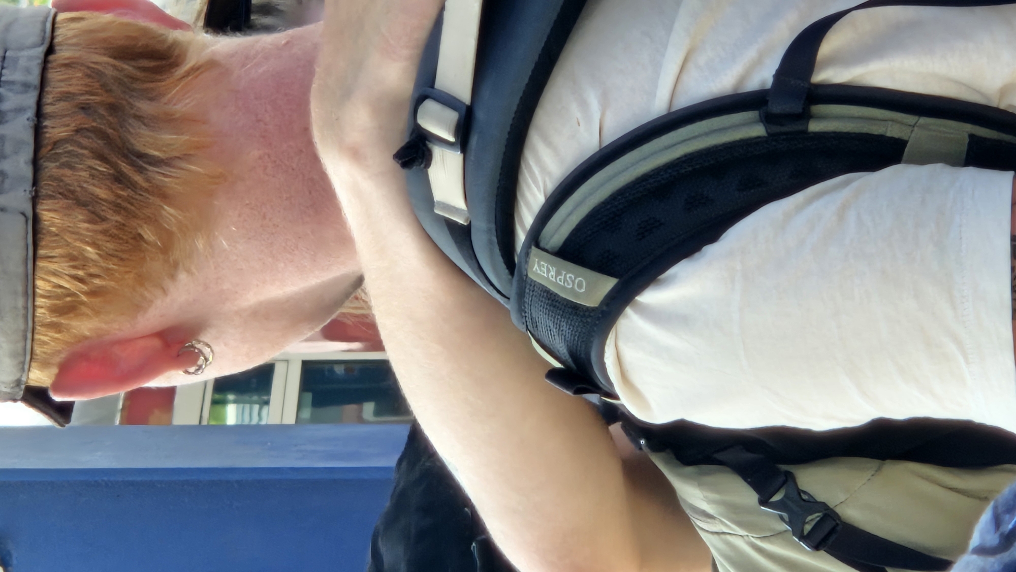 The image shows a close-up view of a person from the back, focusing on their neck, shoulder, and part of their arm. The person has light-colored hair, possibly red or blonde, and is wearing a cap. They have an earring in their left ear, which appears to be a silver hoop. A portion of a backpack is visible, with the brand name "OSPREY" on a tag. The backpack straps are dark and have some grey and green accents. The person is wearing a light-colored t-shirt, and a portion of a tan jacket is also visible. The background is blurred, but a blue structure and some glass or windows can be seen, suggesting an urban or public setting. There is no specific indication of the city of Bratislava, Slovakia, in the image itself.