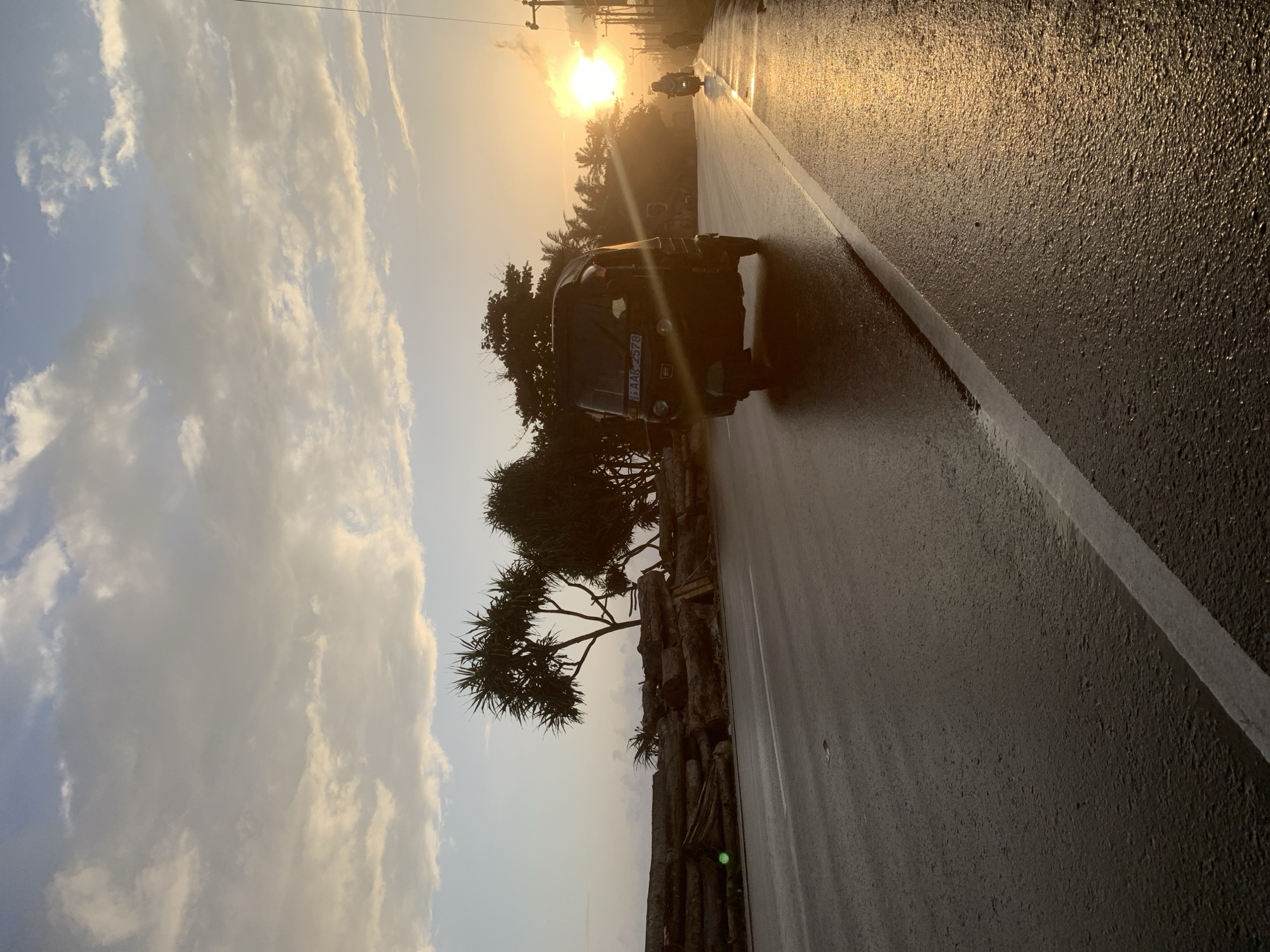 An image taken at sunset on a wet road in Ahangama, Sri Lanka. A dark-colored auto-rickshaw is driving on the right side of the road, reflecting the setting sun. In the distance, a motorcycle with a rider is also visible, along with a few pedestrians. The road is bordered by a pile of logs on the left and palm trees and vegetation on the right. The sky is partly cloudy, with the sun casting a warm glow.