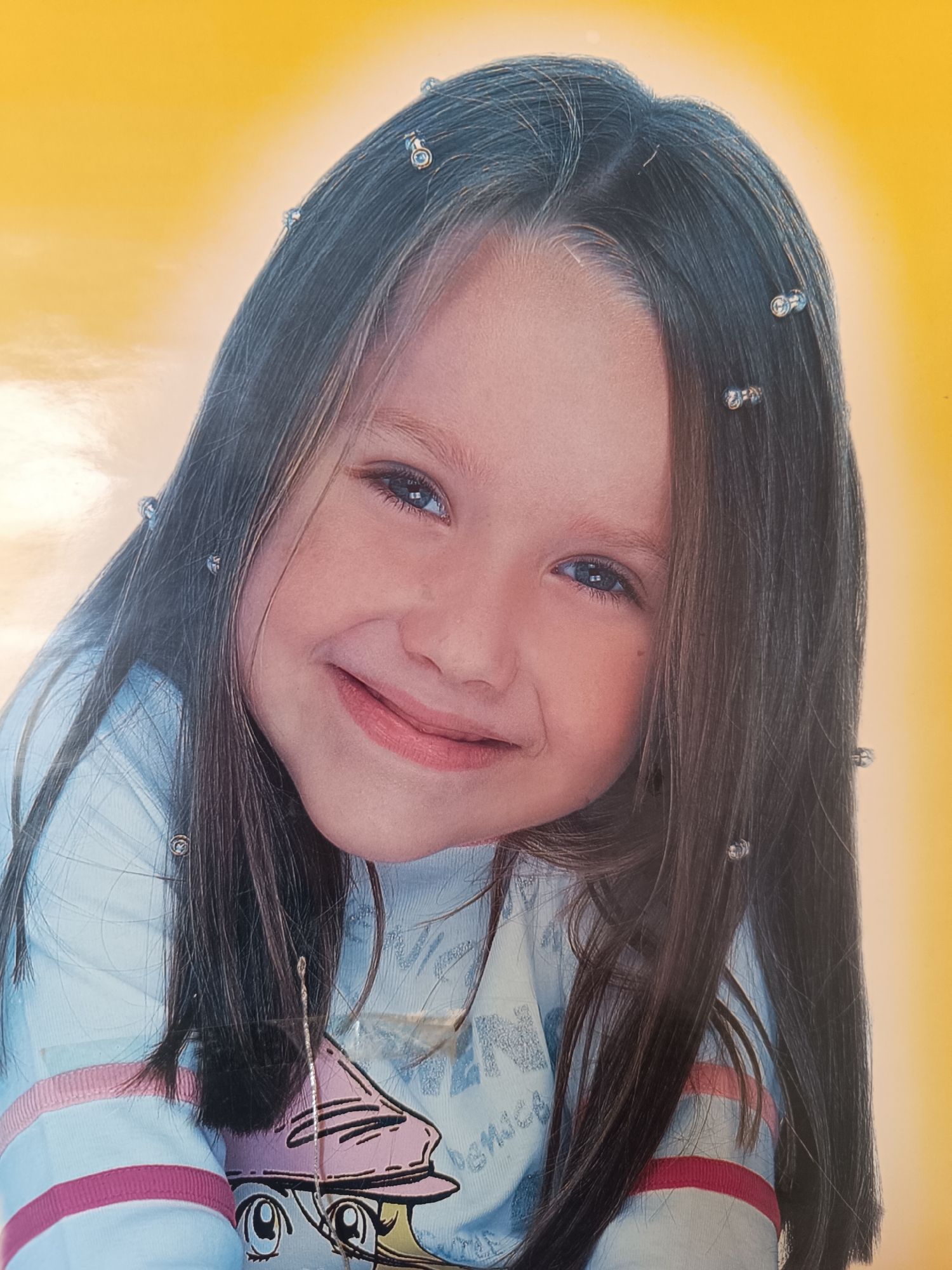 A close-up portrait shows a young girl with brown hair and blue eyes. She is smiling slightly. Her hair is styled with small, round hair accessories. She is wearing a light blue shirt with pink stripes on the sleeves. The background is a gradient of yellow and white. The city is Unknown and the location is Unknown.