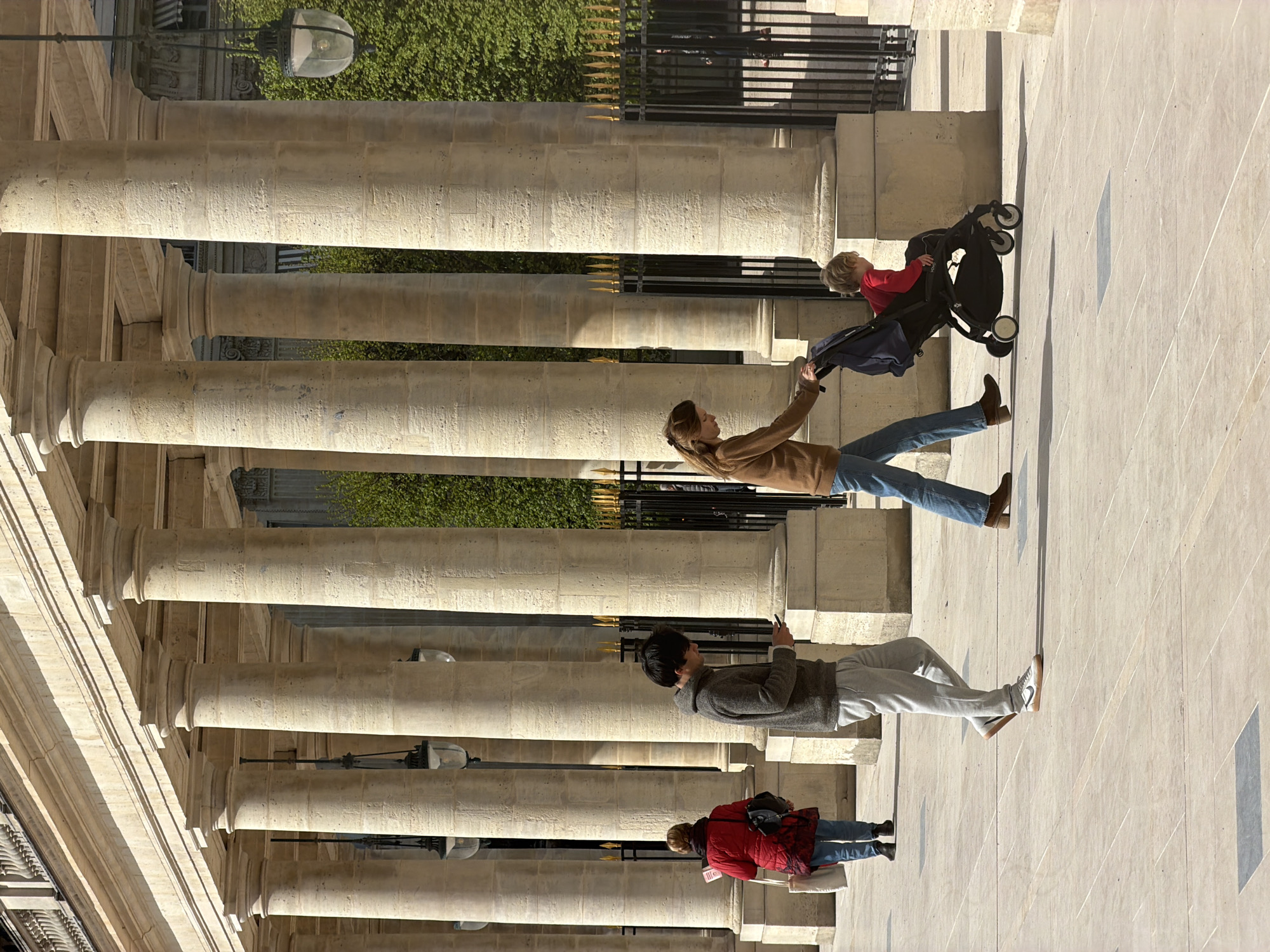 The image shows a row of tan stone columns and a portion of a building’s exterior in an outdoor setting. The people present are interacting with the physical setting, while the woman in the middle is seen pushing a stroller. The building and architectural style suggest this is in Paris, France. The people, columns, sidewalk, and architectural details align in the image to suggest the setting. There are 4 people in the photo, 2 women, one man and one child.