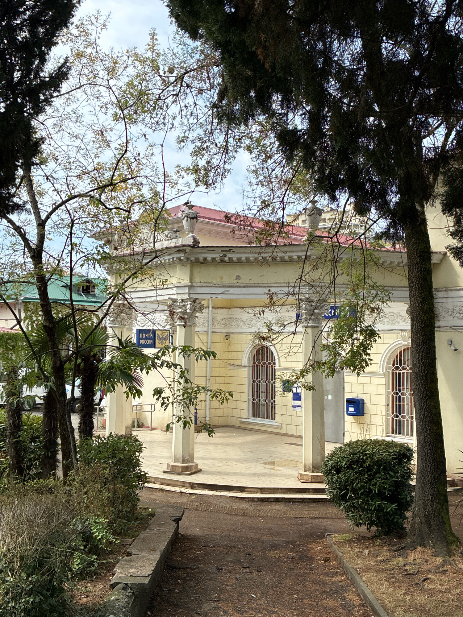 The image shows a view of a post office building with a sign that reads "ПОЧТА РОССИИ" (Russian Post). The building is yellow with white architectural details and a red roof. It has arched windows with decorative grilles and columns supporting an overhang. A blue mailbox is mounted on the wall to the right of the entrance. Palm trees and other foliage are in the foreground, partially obscuring the building. The overall setting appears to be a public space, likely in Sochi, Russia, given the Russian signage. The ground is a dirt path with some scattered leaves. The sky is visible through the branches of trees in the foreground, and it appears overcast.