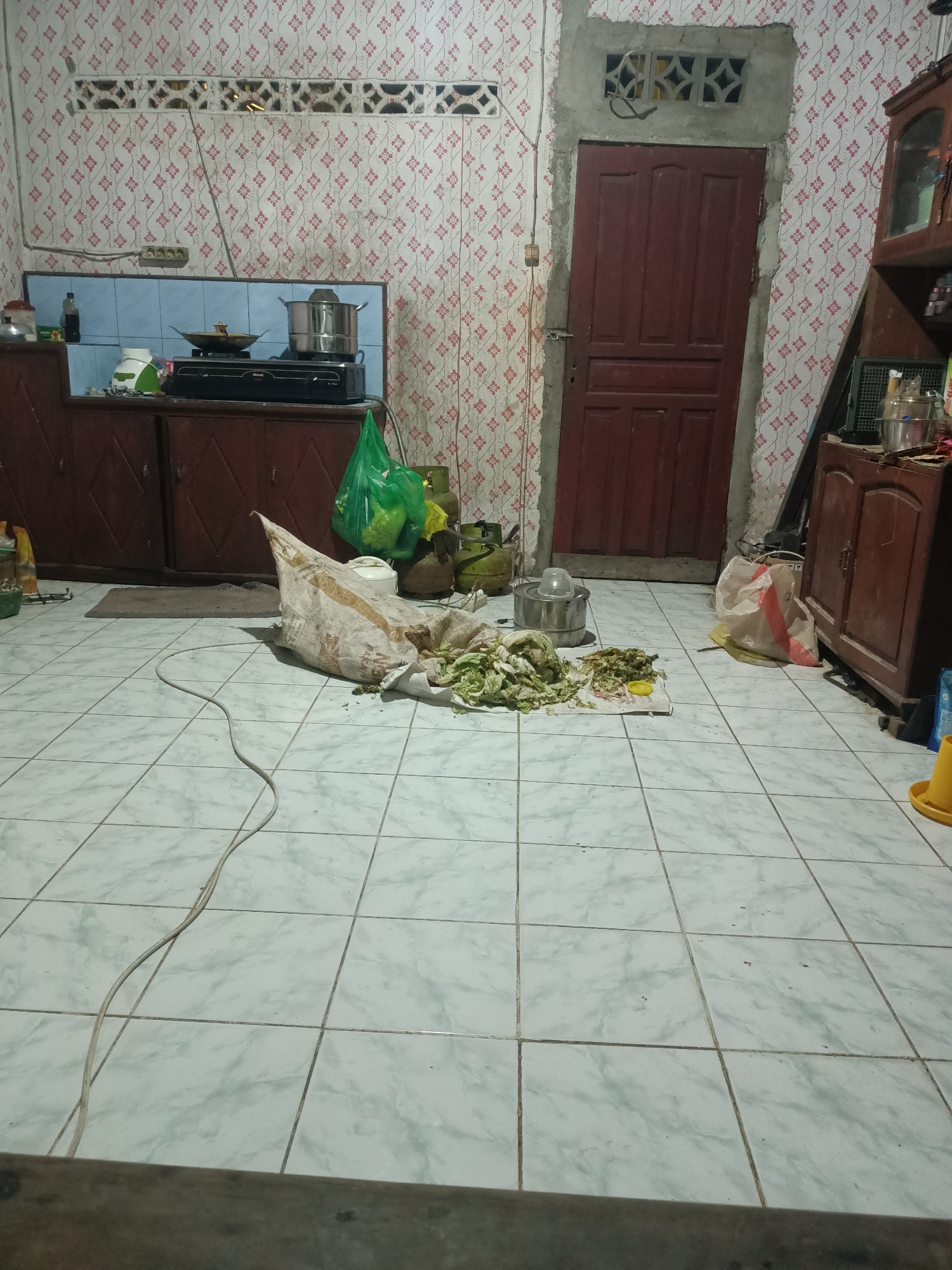 The image depicts an interior scene, likely a kitchen area, within a home in Rasau Jaya Satu, Indonesia. The floor is tiled with light-colored square tiles that have a marbled pattern. On the floor, in the center of the image, is a large, torn sack made of rough material, with chopped vegetables spilling out onto a white cloth. To the left of the sack is a silver pot with a lid, and next to it, a green plastic bag filled with more vegetables. In the background, there is a kitchen counter with a stove, pots, and a bottle. A wooden cabinet with diamond-shaped doors is visible on the left. To the right, a dark red wooden door is partially open, revealing a dark interior. Another wooden cabinet with glass doors is positioned on the far right. The walls are covered with patterned wallpaper featuring small red and white floral designs. A gray electrical cord snakes across the tiled floor. The overall impression is one of a functional, somewhat cluttered domestic space.