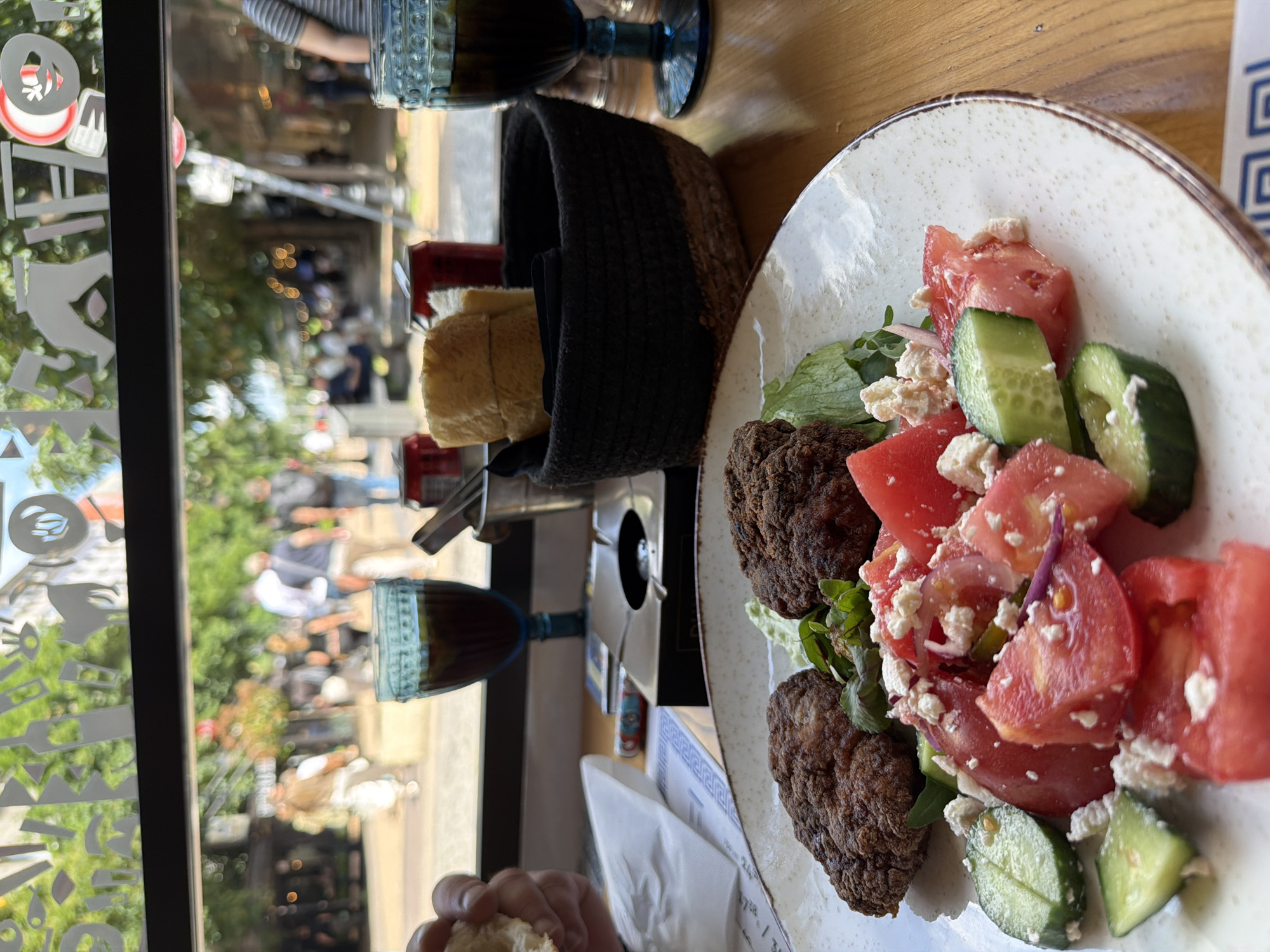 The image depicts a meal at a restaurant in Sofia, Bulgaria. A white plate containing a salad with tomatoes, cucumbers, and what appears to be feta cheese sits next to two dark brown fried meatballs or falafel. A basket filled with bread is nearby. Two blue glasses are also visible on the table. Through a window, a street scene with people walking and sitting is visible. The window has decorative etched patterns.
