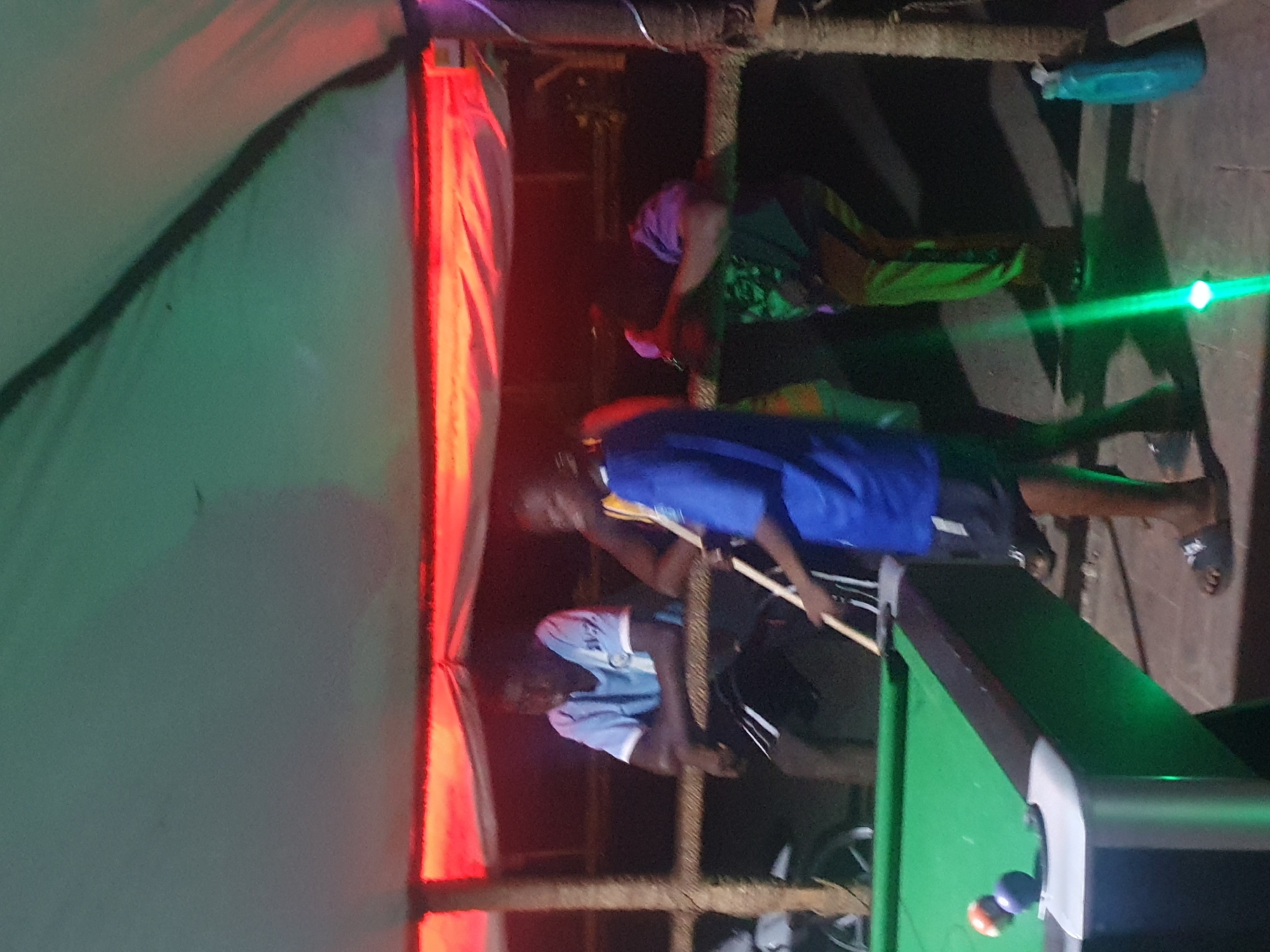 The image depicts an outdoor billiard scene in Lokoja, Nigeria. Two men are playing pool under a makeshift shelter constructed with wooden poles and tarpaulin. The lighting is provided by string lights, casting a red glow on parts of the shelter. One man, wearing a blue shirt, is in the process of taking a shot with a cue stick. Another man, wearing a light blue and white shirt, leans against the side of the pool table, observing. An older woman sits behind them, watching the game. A water bottle sits on one of the poles holding up the shelter. The background is dark, suggesting it is nighttime. A green laser beam is also visible in the shot.