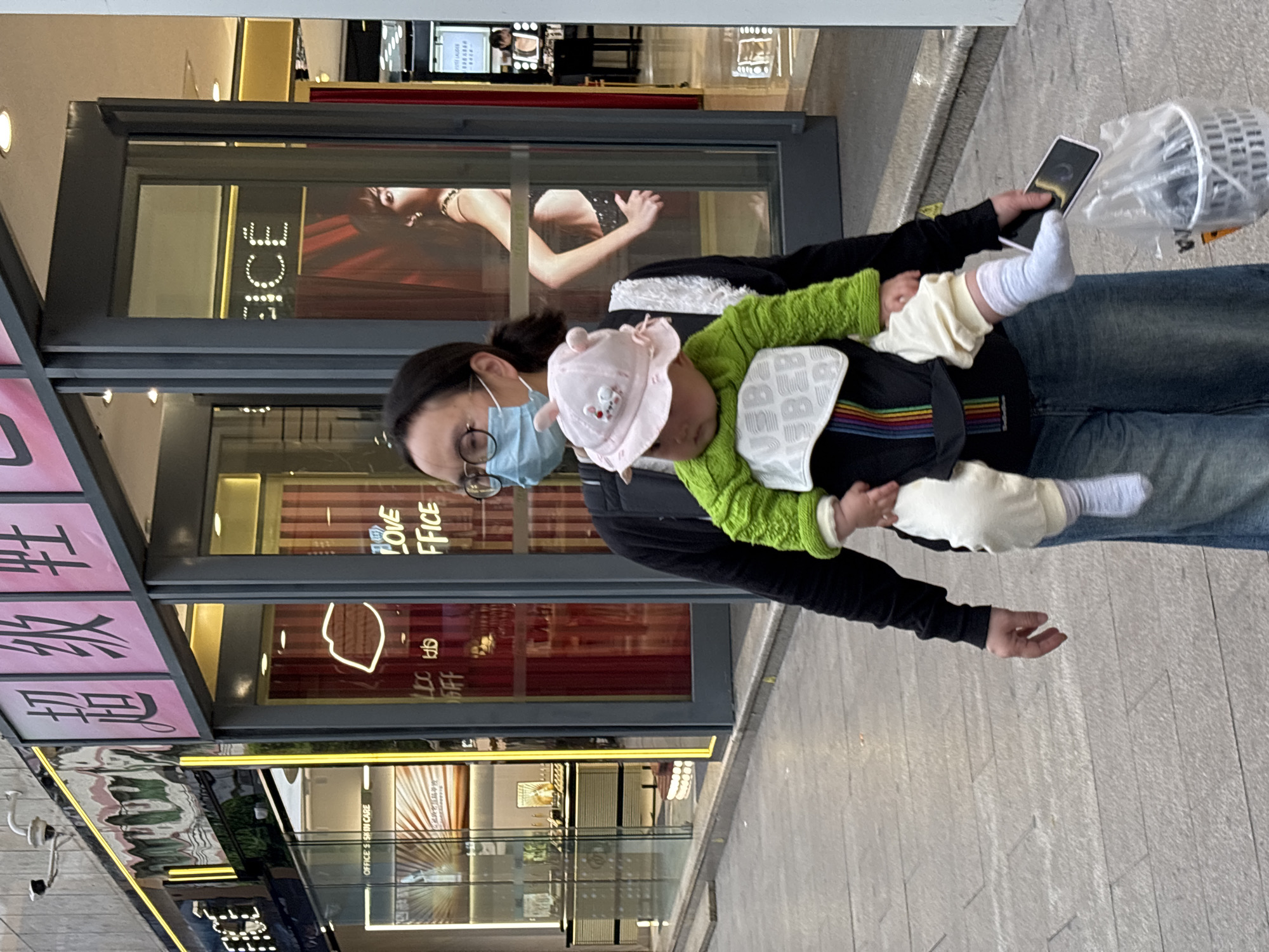 A medium, street-level shot captures a person carrying a baby in front of storefronts, possibly located in Jiangjin, China. The person, likely a parent, wears glasses and a blue face mask and is dressed in a black long-sleeved shirt and denim jeans. They are holding a smartphone in one hand and carrying a shopping bag. The baby, secured in a baby carrier, is wearing a light green sweater, white bib, and a pink hat. The backdrop consists of commercial storefronts, with the storefronts appearing to have reflective windows. The location is implied by the Chinese characters on the exterior of the storefront. The ground consists of horizontally aligned tiles of a light grey color.
