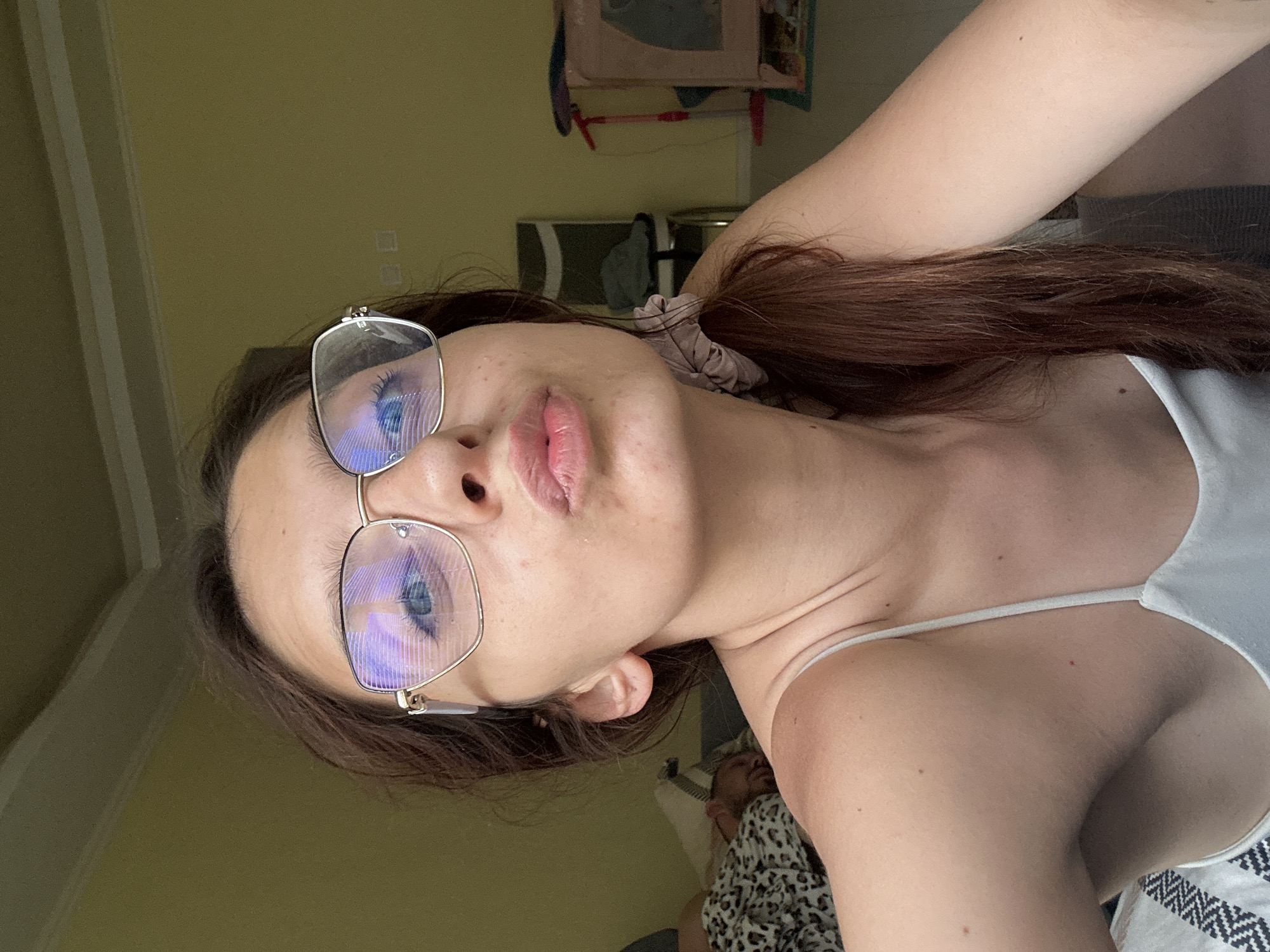 This is a close-up, vertically oriented selfie of a young woman indoors. She is wearing glasses with thin, metallic frames and has her hair pulled back with a scrunchie. Her lips are slightly parted, and she appears to be looking at the camera. The background is softly lit with a yellowish-green hue, suggesting it might be daytime with natural light coming from a window. There are some blurry objects in the background, including what looks like shelves and possibly some framed pictures or decorations. The woman's right arm is visible, resting behind her head. The overall mood is casual and relaxed. There is no visible text in the image. Given the location context of Tryavna, Bulgaria, this could be a personal snapshot taken in a home environment.