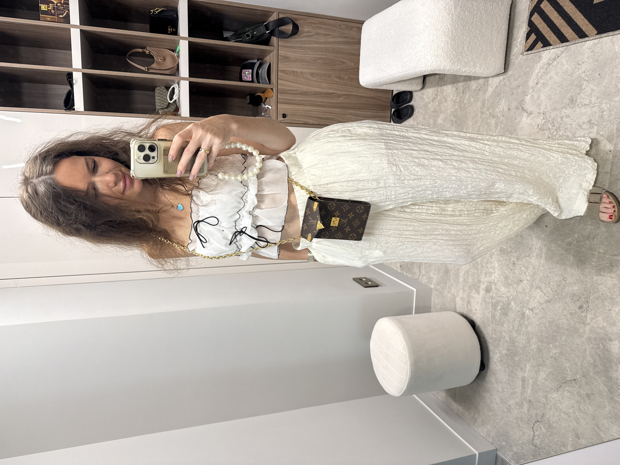A woman is taking a selfie in what appears to be a walk-in closet or dressing room. She is wearing a white, ruffled top, a long white skirt or pants with a textured pattern, and accessorized with a gold chain bag and a pearl necklace. She is holding her phone up to take the picture. The room has built-in shelving with various items, including handbags and accessories, neatly arranged. A fluffy white ottoman is visible on the right side of the frame, and a smaller round ottoman is positioned near the bottom of the frame. The floor is a light grey marble or tile. The scene does not provide information to determine the specific location in Moscow, Russia.