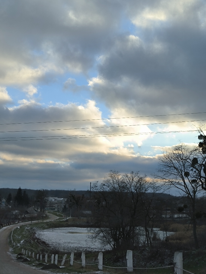 An elevated view captures a wintry landscape in Khrinivka, Ukraine, under a dramatic sky. The foreground features a dirt or gravel road, lightly covered in patchy snow or frost, curving leftwards and descending out of frame. Along the right edge of this road, a low fence composed of regularly spaced white posts connected by a horizontal rope or wire is visible, also dusted with snow. To the right of the road and fence, a partially frozen pond, its surface a dull white, is surrounded by bare deciduous trees and shrubs, some with traces of snow.
In the midground, the road continues to curve, leading towards a cluster of buildings interspersed with trees. Several multi-story structures, possibly residential or institutional, are visible amongst the leafless trees. A tall, slender industrial-style chimney or vent stack rises on the left side of the midground. The background consists of a rolling, tree-covered landscape, with additional scattered buildings.
The sky is heavily overcast with prominent grey and white cumulus clouds. Patches of brighter blue sky are visible through breaks in the cloud cover, particularly towards the upper center and right. Sunlight, possibly from a low-lying sun, illuminates the undersides and edges of some clouds, creating highlights. Multiple utility wires stretch horizontally across the upper portion of the frame. No individuals or active subjects are present in the scene.