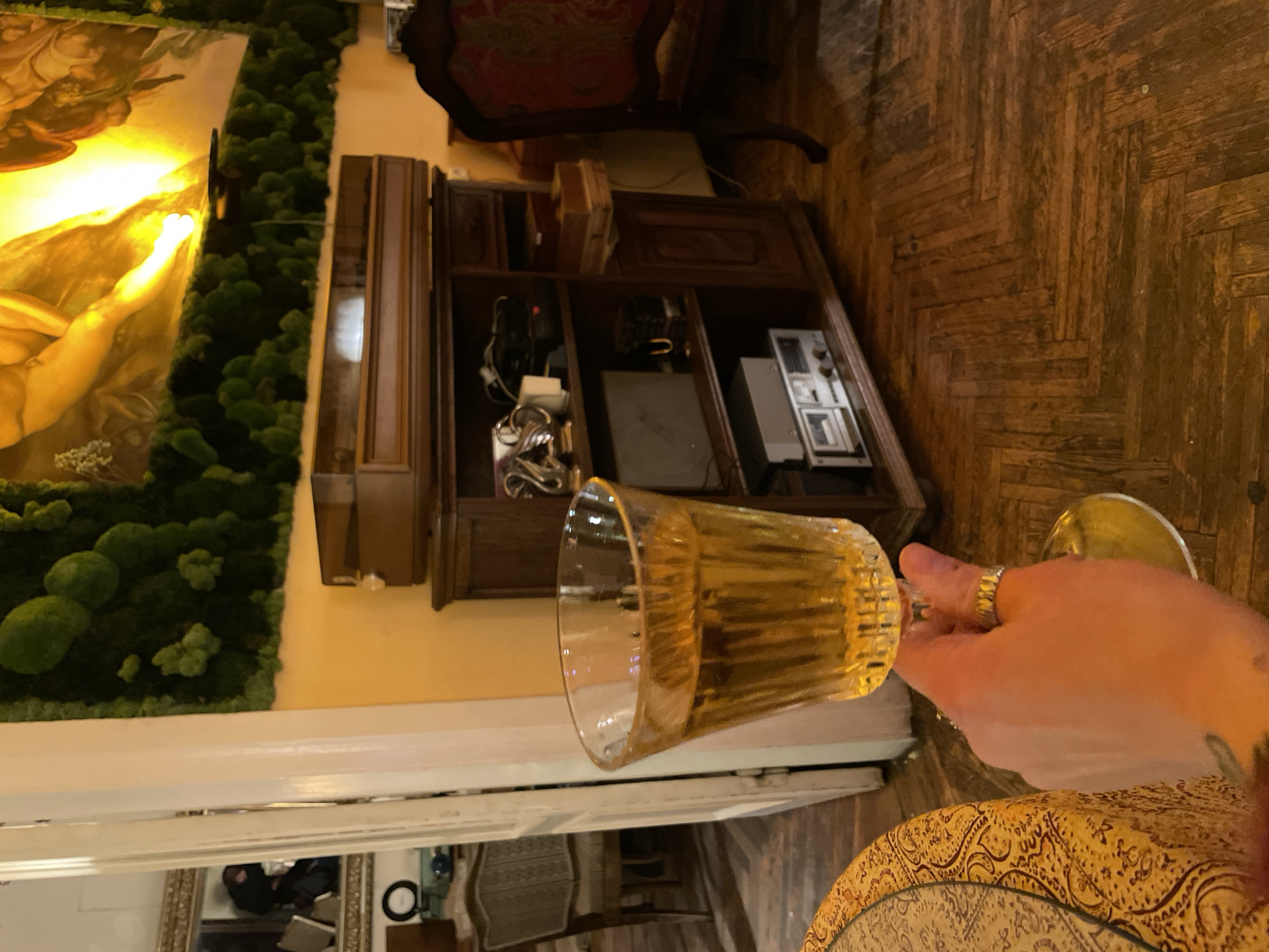 A tilted, close-up indoor scene features a hand holding a partially filled glass of a golden liquid, likely a beverage, against a backdrop of mixed architectural and decorative elements. The hand, adorned with a ring, seems to be in the act of offering or displaying the drink.
Behind the glass, a wooden cabinet holds various items, including what appears to be vintage electronic equipment. To the left, a section of wall art, possibly a painting, is framed by a vertical arrangement of moss-like greenery.
A glimpse into another room is visible through a doorway in the background, suggesting a residential or hospitality setting. The image has an overall warm tone, indicative of the lighting and the color palette of the environment.
The city of Tbilisi, Georgia is not discernible from the details in the image.