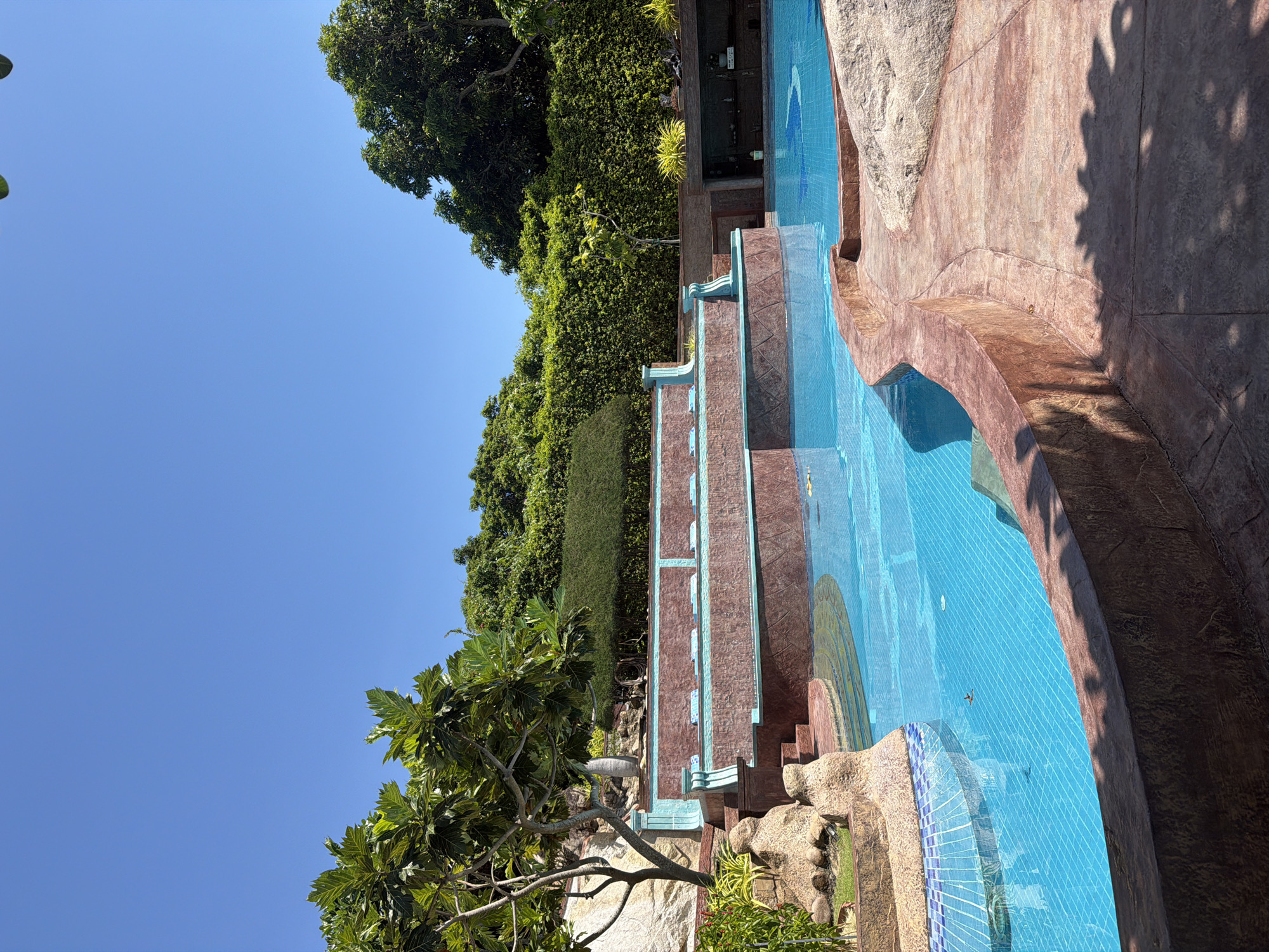 The image shows a tiered swimming pool with blue tiles and artificial rock features, located in Bac Nha Trang Ward, Vietnam. The pool is surrounded by lush greenery, including trees and manicured hedges. A clear blue sky is visible above. There are no people visible in the pool or immediate surroundings. The pool's design incorporates brown and turquoise accents. Shadow patterns suggest a sunny day.