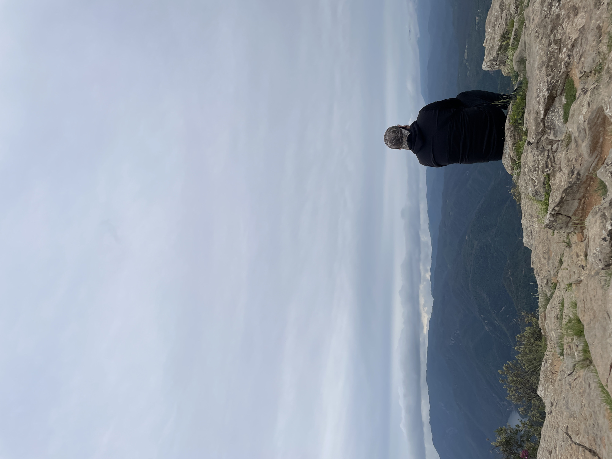 A man is seen sitting on the edge of a rocky cliff, overlooking a mountainous landscape in Tavertet, Spain. He is wearing a dark-colored hooded garment and a camouflage cap. The sky is overcast, and the vista stretches across layered mountains, with some vegetation growing on the cliff's edge.
