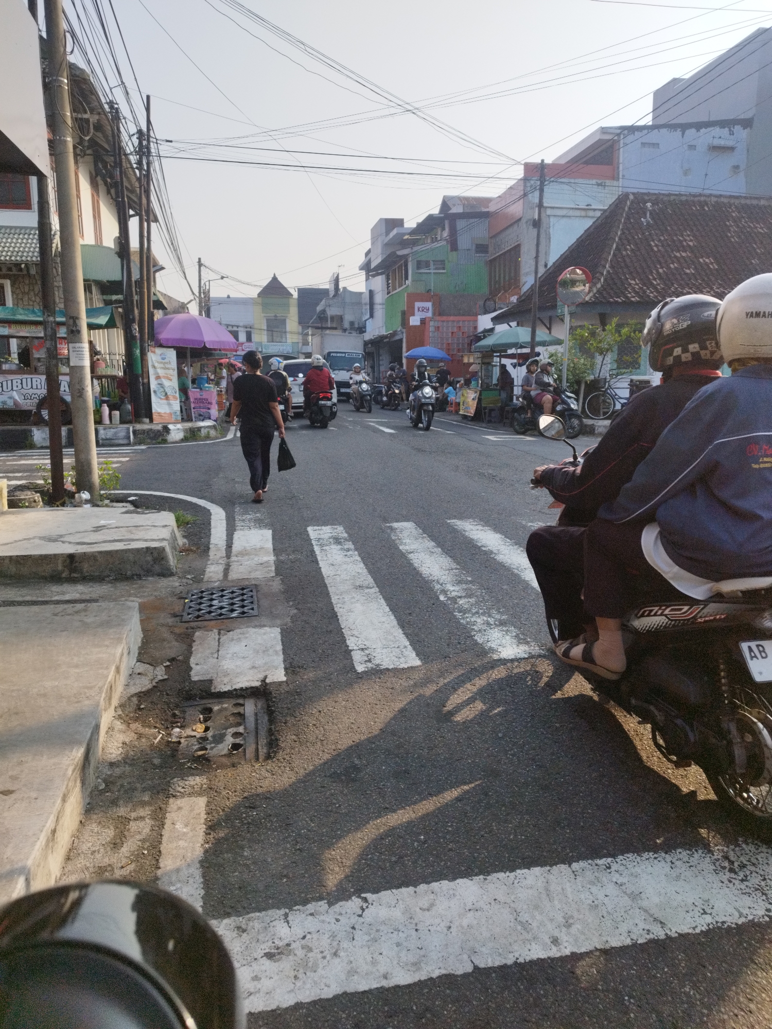 The image depicts a street scene in Yogyakarta, Indonesia, likely during the late afternoon based on the lighting and shadows. A pedestrian in dark clothing walks across a crosswalk on the right side of the image, carrying a black bag. Two people on a motorcycle, wearing helmets, are stopped at the crosswalk, with the passenger in the rear wearing a blue jacket. In the background, several other motorcycles and a truck are visible on the road. Shops and buildings line the street, with various signs and colorful awnings. Overhead, a network of electrical wires crisscrosses the sky. The road surface is asphalt, marked with white pedestrian crossing lines and a metal drainage grate on the sidewalk. The overall environment suggests a busy urban street with moderate traffic.