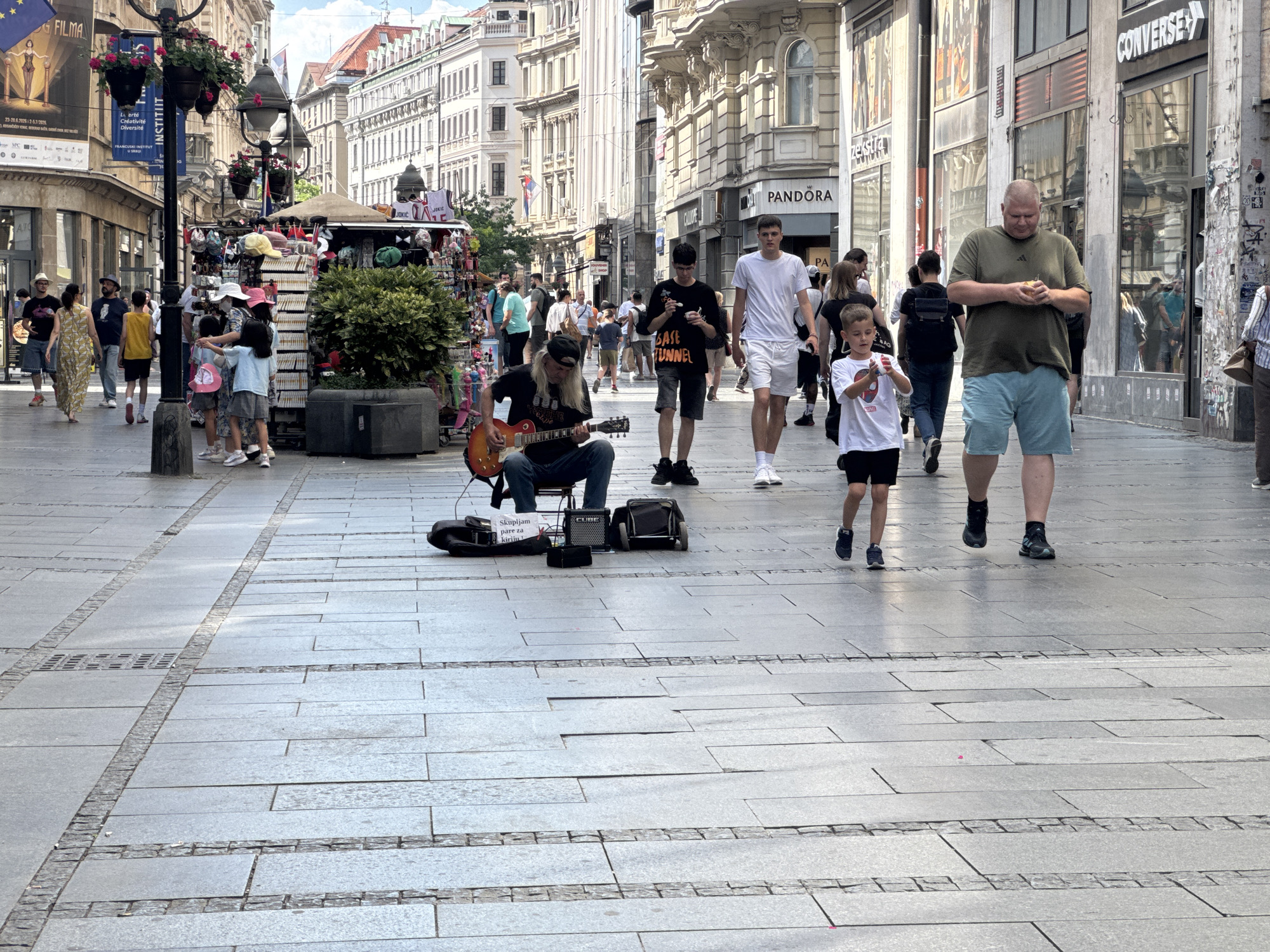 This media file shows a bustling pedestrian street scene in Belgrade, Serbia. The time of day appears to be midday, with bright sunlight casting distinct shadows on the paved walkway. The weather seems pleasant, contributing to the lively atmosphere.
The main subjects are various people walking along the street, including families, couples, and individuals. In the foreground, a street musician is playing an electric guitar while sitting on a stool. His guitar case and a small amplifier are placed on the ground in front of him, and a sign that reads "Skupljam pare za kiriju" (I'm collecting money for rent) is visible.
Other notable elements include several shops lining the street, with prominent signage such as "PANDORA" and "CONVERSE" visible. There are also market stalls selling various items, including hats and souvenirs. The architecture of the buildings suggests a European city setting, with ornate facades and multiple stories.
The overall mood is one of everyday urban activity and a sense of community, with people enjoying a walk in the city center. The diverse crowd and the presence of a street performer create a vibrant and engaging scene.