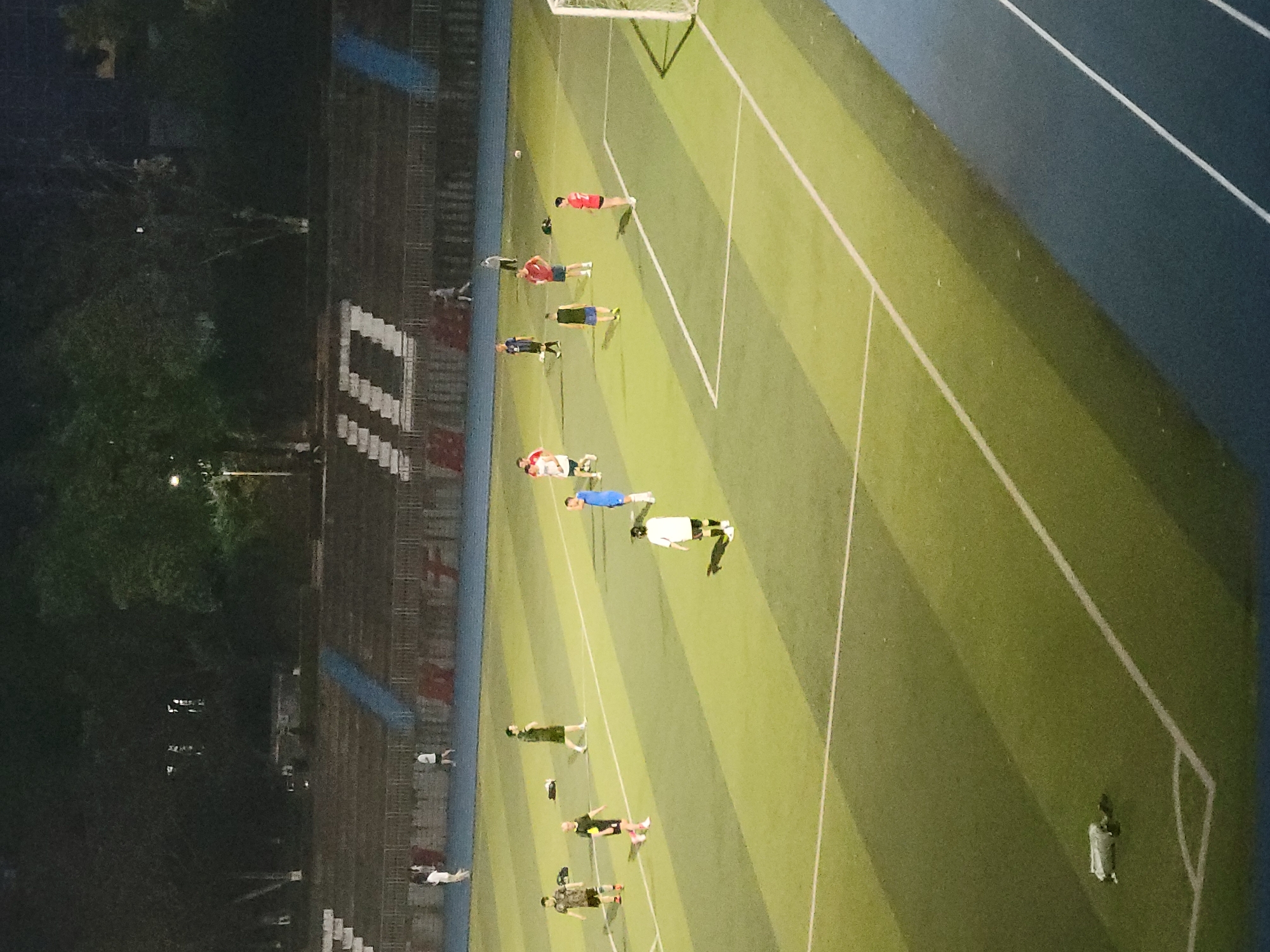 The image shows a soccer field in Shapingba District, China. Several people are scattered across the field, some appearing to be engaged in stretching or warm-up exercises. The field is well-lit. A track surrounds the field on one side. A small set of bleachers is visible along one side of the field. The lighting conditions suggest it is nighttime.