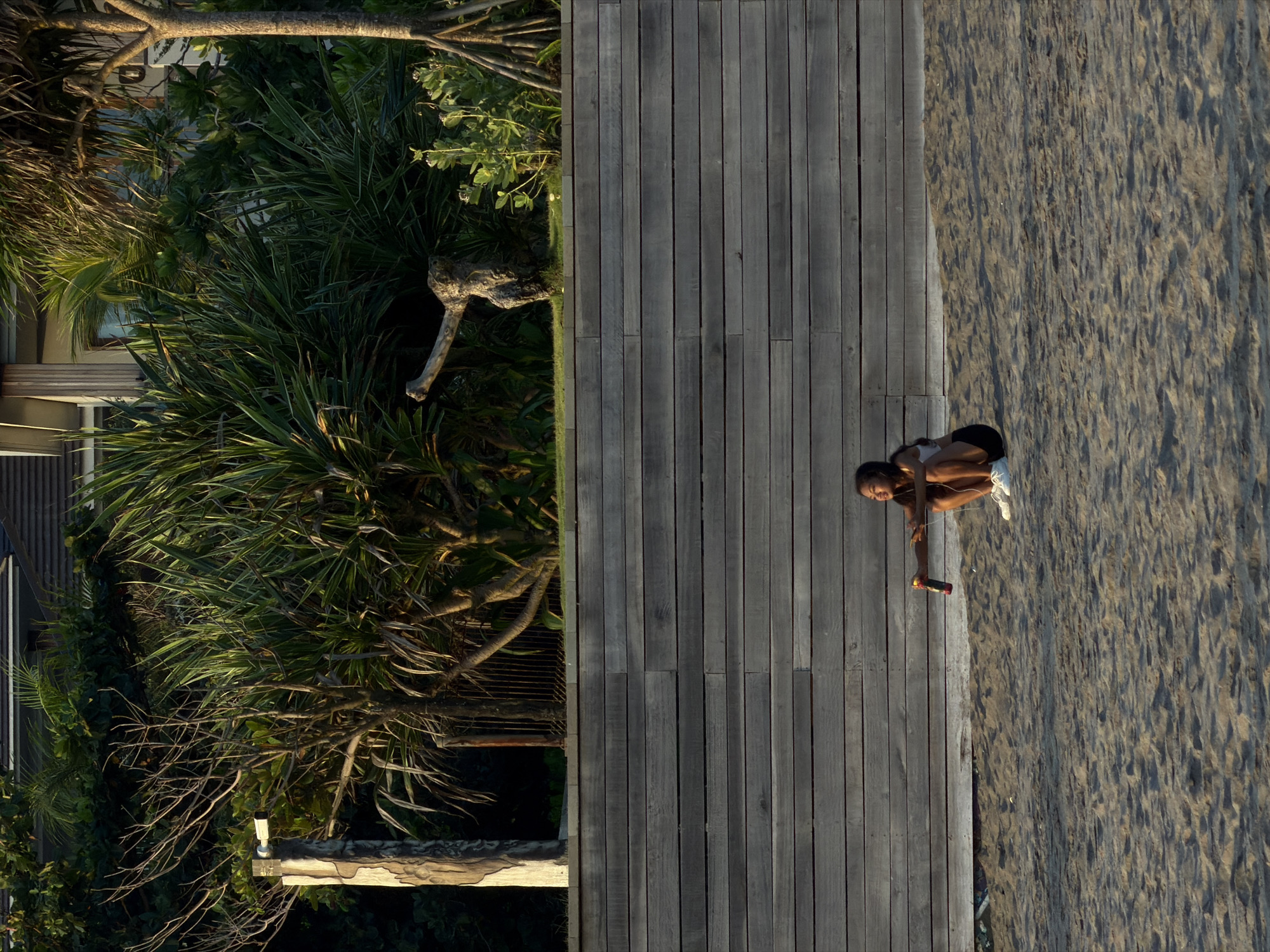 A young woman with dark hair is sitting on a sandy beach in North Kuta, Indonesia. She is wearing a white tank top, black shorts, and white sneakers. She is holding a bottle in her right hand and appears to be looking to her left. The beach is backed by a horizontal wooden wall, and behind that is a lush green area with palm trees and vegetation. The sun is casting long shadows, indicating it is either early morning or late afternoon.