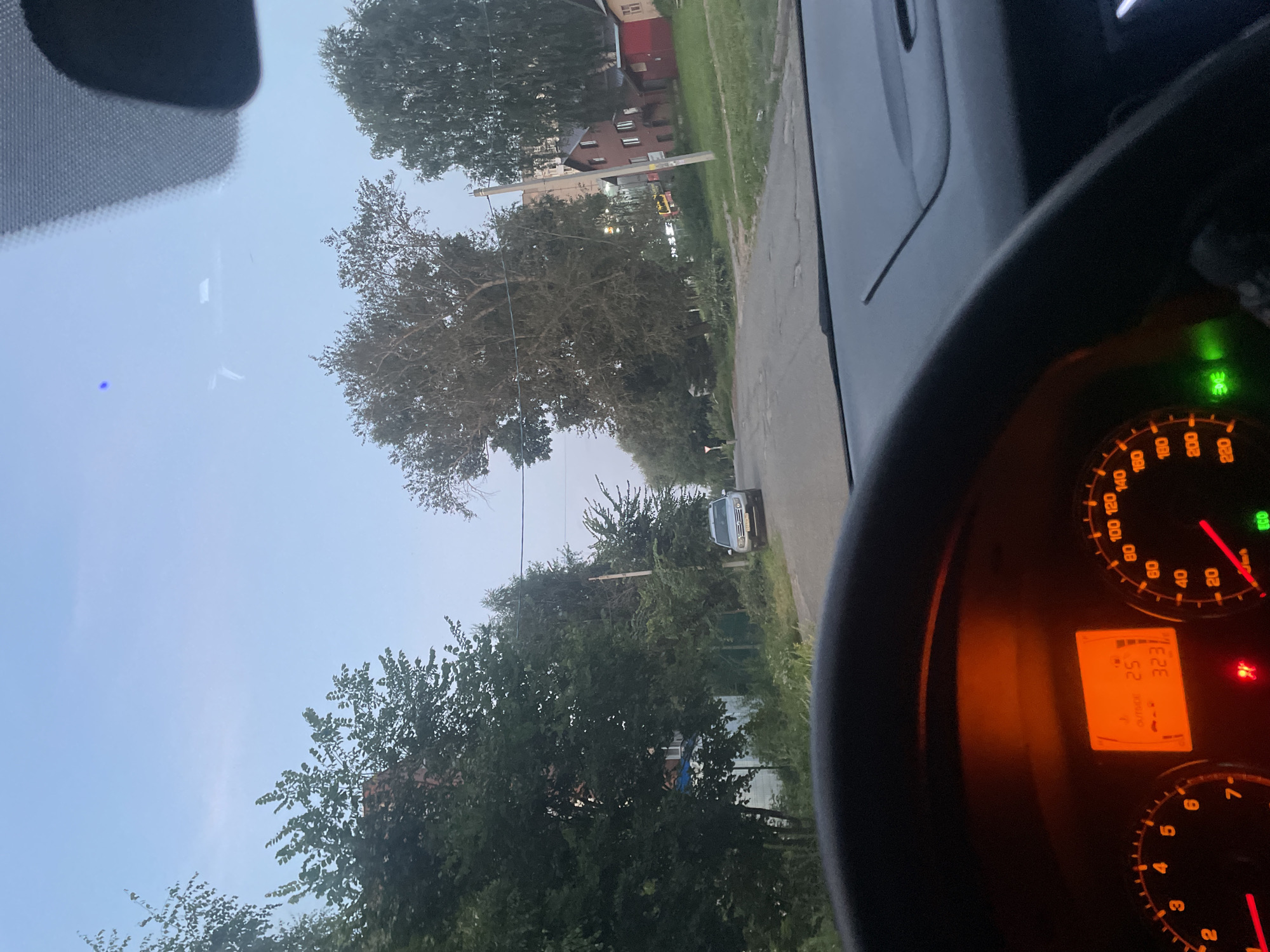 The image, captured from the driver's seat of a car, depicts a street scene in Votkinsk, Russia. The sky is a pale blue with a few wispy clouds. The road ahead is paved and appears to be in moderate condition with some visible cracks. Trees and houses line the street, with a mix of deciduous and coniferous varieties. In the mid-ground, a silver SUV is parked on the right side of the road. A multi-story building and a smaller red house are visible behind the trees on the right. The car's dashboard is illuminated, showing the speedometer and a digital display indicating an outside temperature of 25 degrees Celsius and a time of 3:23. The steering wheel is in the foreground, partially obscuring the dashboard. There are no people visible in the image.