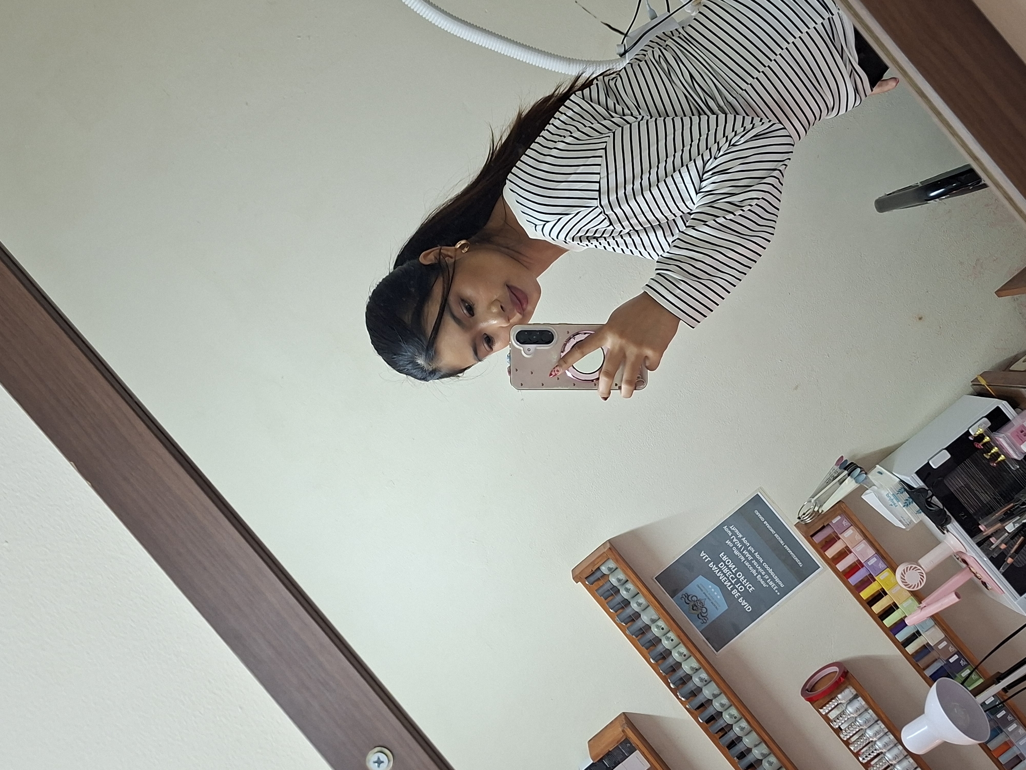 In a beauty salon in Kuta, Indonesia, a young woman with dark hair pulled into a ponytail is taking a mirror selfie. She is wearing a black and white striped long-sleeved shirt and gold hoop earrings. She is holding a pink phone with a decorative case in her right hand, which is positioned to capture the reflection. The background shows shelves filled with nail polish bottles, a small desk fan, a lamp, and what appears to be a UV curing lamp. A sign with text is visible on the wall. The salon appears to be well-lit.