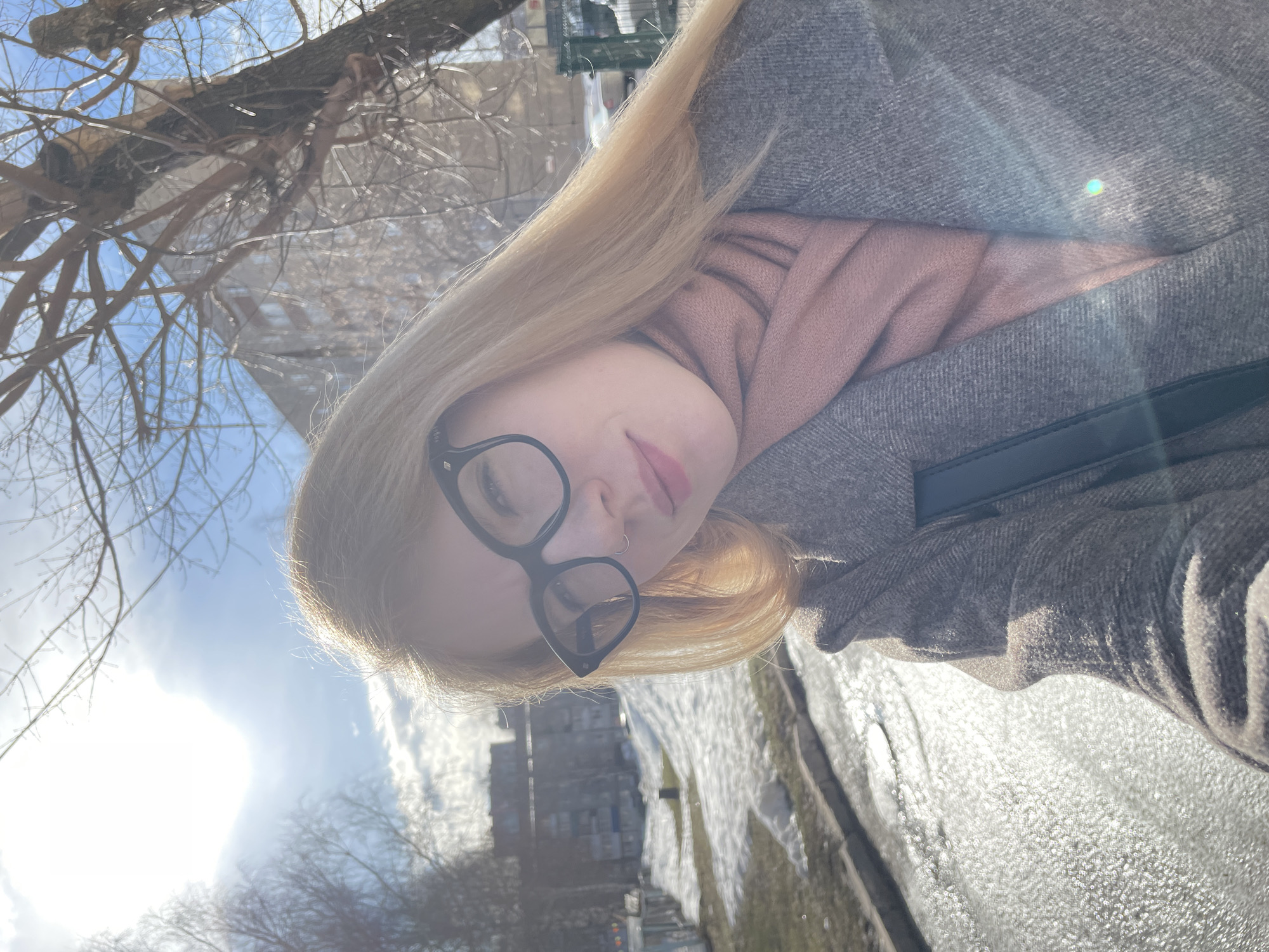 This image depicts a female individual in a residential area of Izhevsk, Russia, captured during daylight hours. The subject occupies the right and central portions of the frame, facing the camera. She has long, straight, blonde hair, wears black-framed eyeglasses, and a silver ring in her left nostril. Her attire includes a gray, textured overcoat and a pink scarf wrapped around her neck. A black strap, consistent with a bag strap, is visible over her left shoulder under the coat's lapel. The subject's mouth exhibits a slight upturn.
The background shows a bright, blue sky with scattered white clouds. Intense sunlight from the upper left quadrant creates a prominent lens flare. Bare deciduous trees are visible throughout the scene, with a large, gnarled tree trunk present on the right side. The ground consists of uneven patches of melting snow and ice, intermingled with wet, reflective surfaces indicative of a street or paved area. A concrete curb runs along the left side of the frame. Multiple multi-story residential buildings, primarily gray or light-colored, are discernible in the mid-ground and distant background, consistent with Izhevsk's urban development. The presence of snow melt and bare trees suggests a late winter or early spring season.
