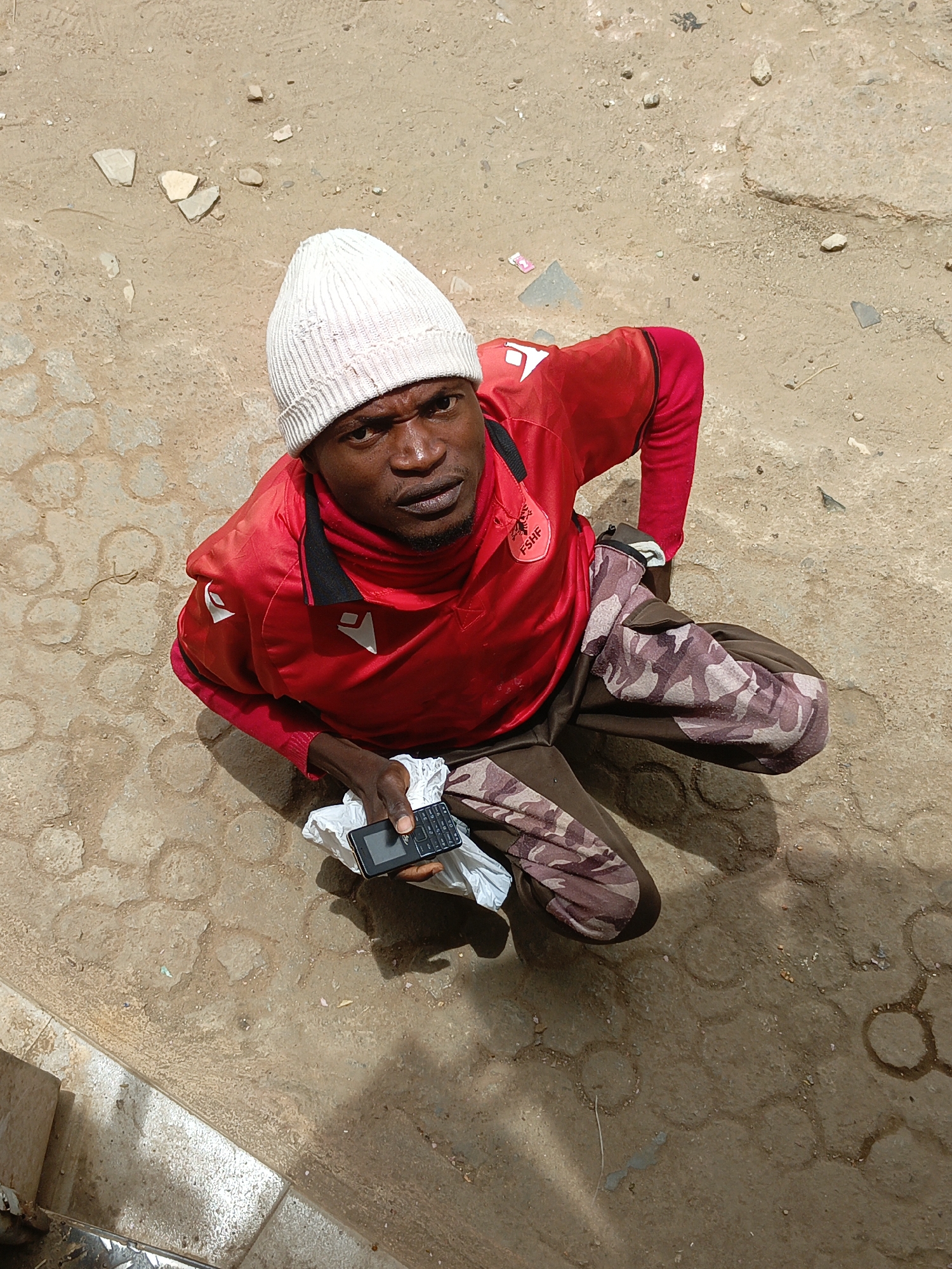 A man wearing a white beanie, a red long-sleeved shirt, and camouflage patterned pants is kneeling on a paved surface outdoors. He is holding a black mobile phone in his left hand and is looking upwards toward the camera. The ground is a mix of light brown and beige with visible paving patterns. The scene appears to be in Jos, Nigeria.
