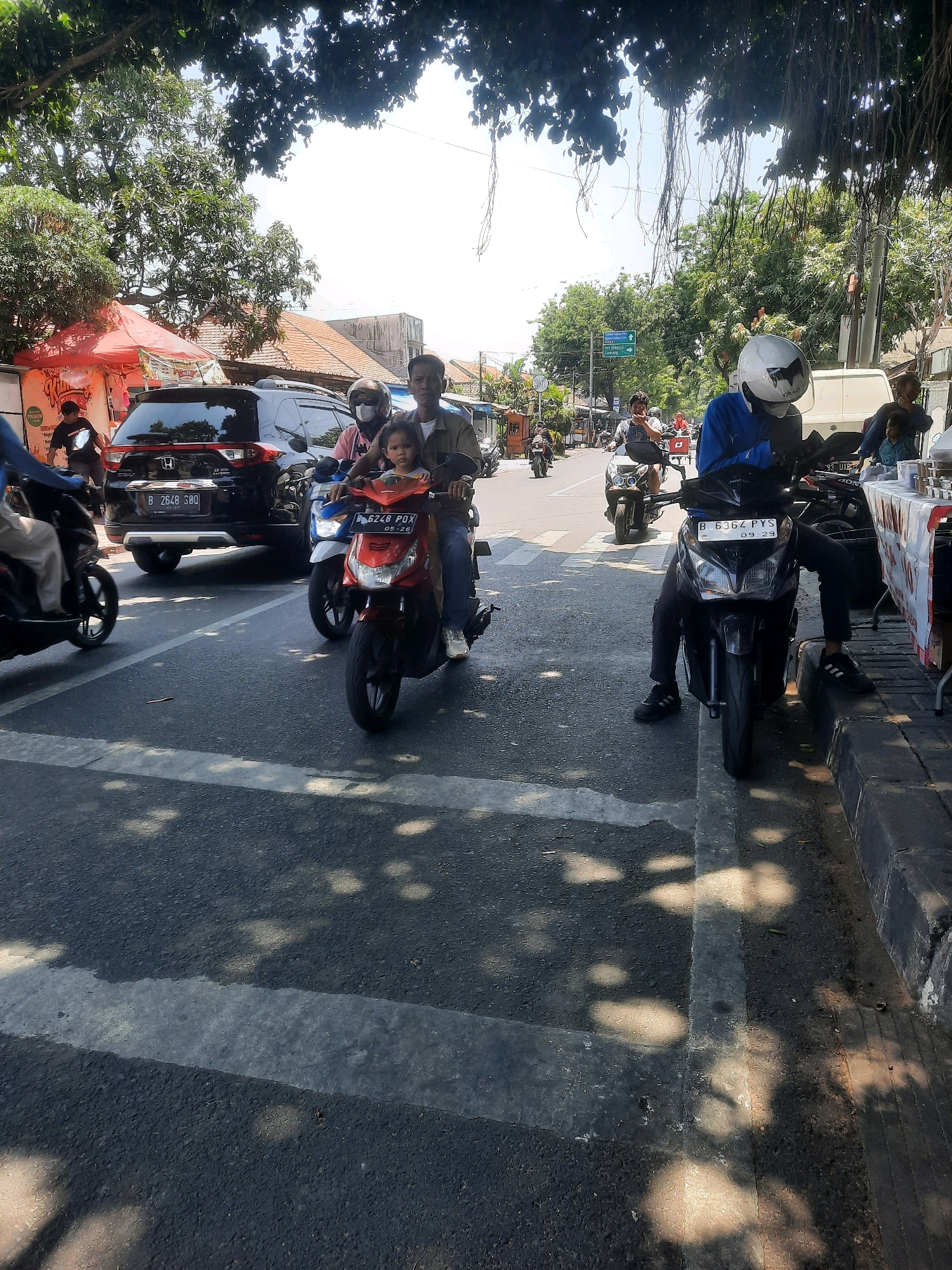 A daytime street scene in North Jakarta, Indonesia, shows several motorcycles on a road, with a black SUV parked nearby. A red tent is set up on the left side of the road, and a white van is visible in the background. Sunlight casts dappled shadows on the road, suggesting the presence of overhead trees. People are riding on the motorcycles, some wearing helmets. The road is paved, and a pedestrian crossing is marked with white stripes. Buildings and trees line the street.