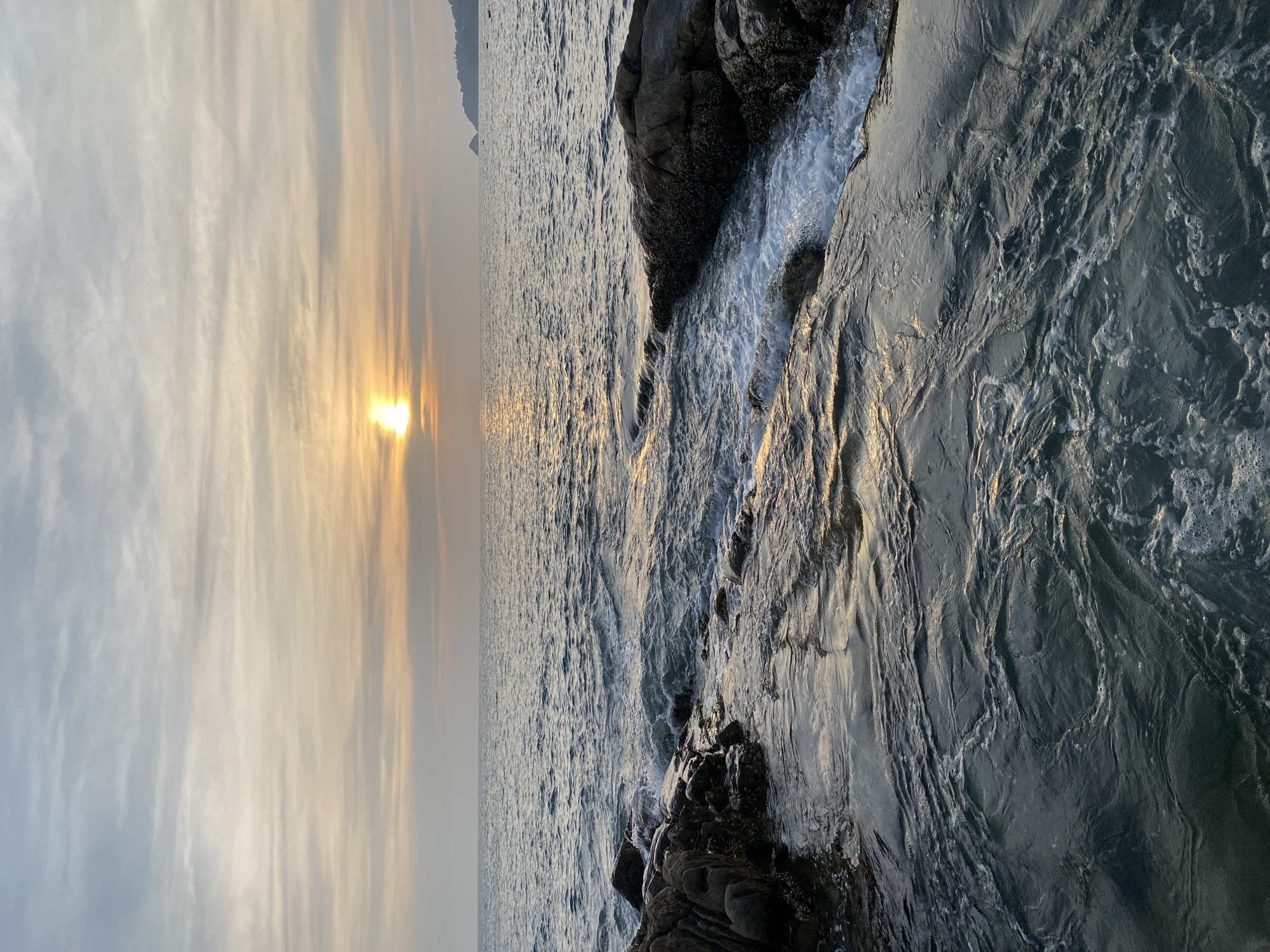 The image shows a sunset scene at the coast. The sun is setting over a body of water, reflecting light on the surface. The sky displays layered cloud formations with colors ranging from gray to orange. The coastline consists of rocky formations being washed by waves. The photograph appears to be taken in Patong, Thailand, though it is unconfirmed if people are present in the image.