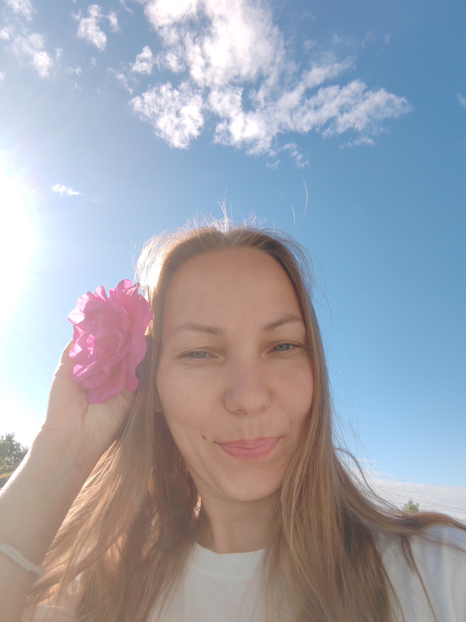 A woman is taking a selfie outdoors with a pink flower near her ear. The background is a bright blue sky with some scattered white clouds. The sun is visible on the left side of the image, creating a lens flare effect. The woman has long, light brown hair and is wearing a white t-shirt. The image appears to be taken in a natural outdoor setting, possibly a park or garden. No specific details of the city of Чухлэм, Russia are visible in the image.