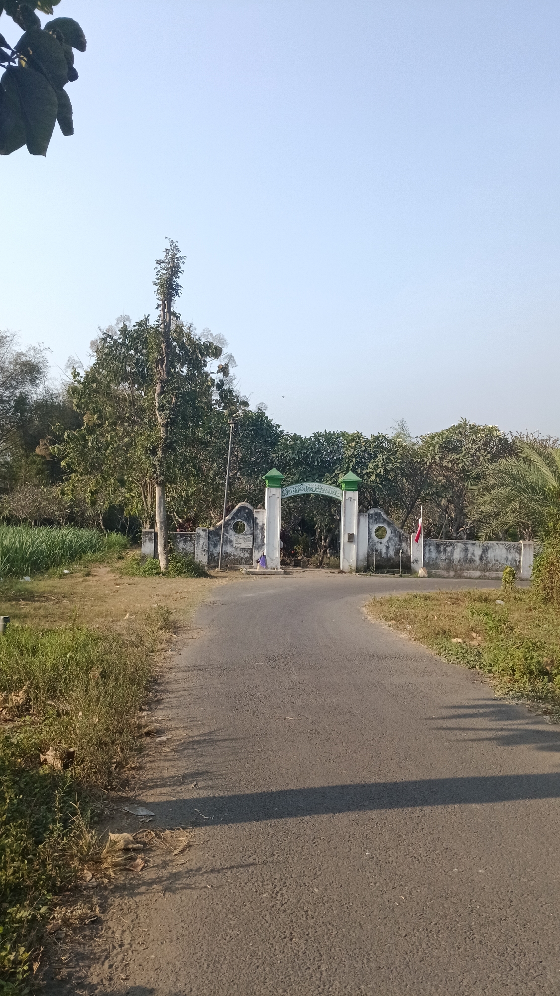 The image depicts an outdoor scene in Slumbung, Indonesia, on a clear day with a pale blue sky. A paved road leads towards an ornate white archway, flanked by low walls and lush green trees. The archway is adorned with Arabic calligraphy in green lettering and features decorative circular openings. To the right of the archway, a small Indonesian flag flies from a pole. The immediate surroundings of the road consist of dry grass, dirt, and fallen leaves. A single person, wearing a dark purple garment, is visible standing near the archway. The overall impression is of a tranquil, rural entrance to a location likely of cultural or religious significance.