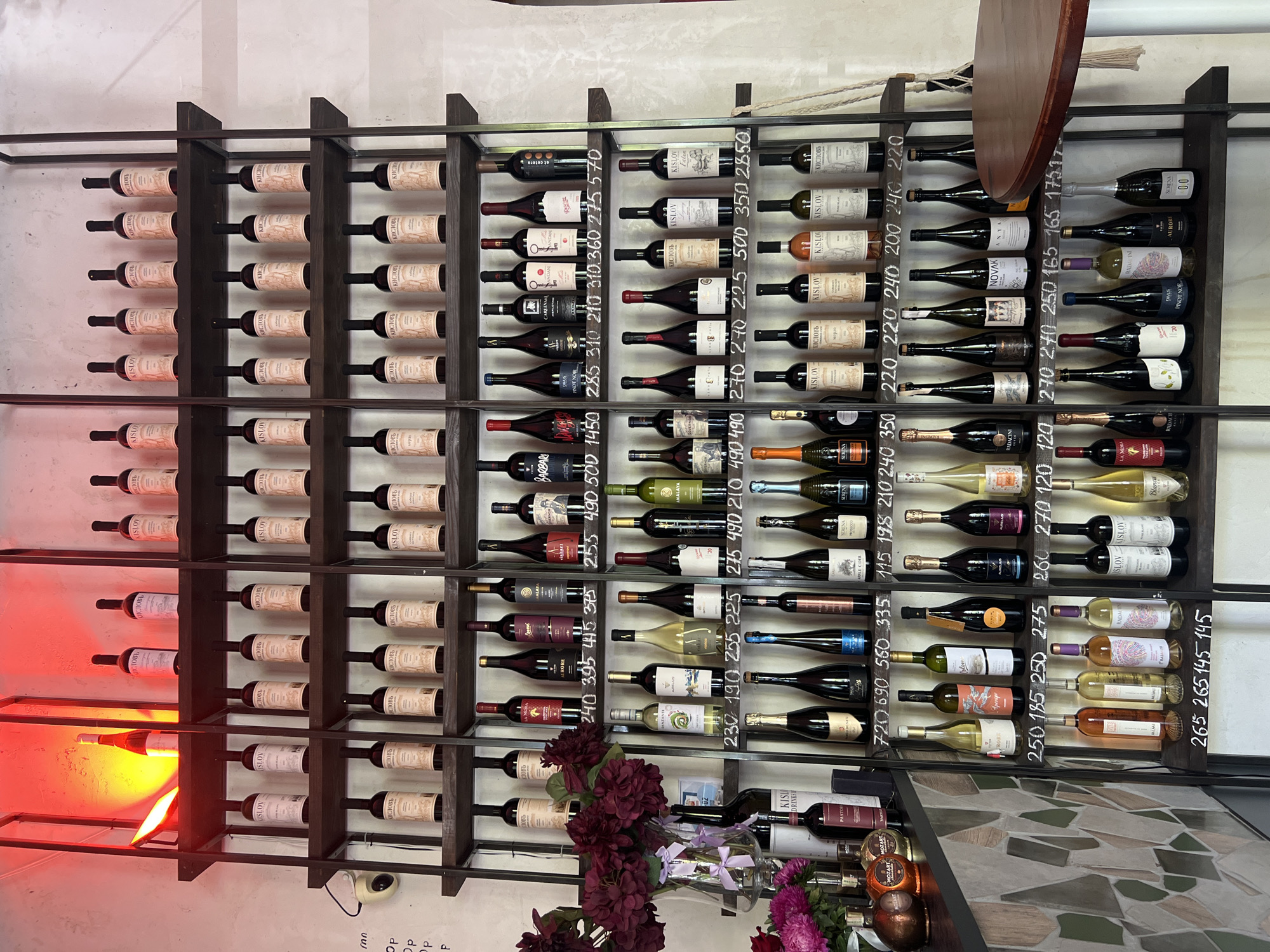 The image shows a wine shelf in a dimly lit establishment. There are multiple rows of wine bottles, most of which are of similar shape and color. The shelves are made of dark metal, and the background is a textured, light-colored wall. Some of the bottles have labels with Cyrillic script. There are also price tags visible below some of the bottles, indicating costs in local currency. The lighting in the upper section of the shelf is a bright red, suggesting it might be a decorative element or a heat lamp. A round, dark-colored table is visible on the right side of the image. A security camera is mounted on the wall to the left. The overall impression is of a wine shop or a bar. The style of the labels and the presence of Cyrillic script suggest the location is in a region where this language is spoken. Based on the visual cues, this establishment is likely in Bender, Moldova, a city with a significant Russian-speaking population and known for its wine culture.
