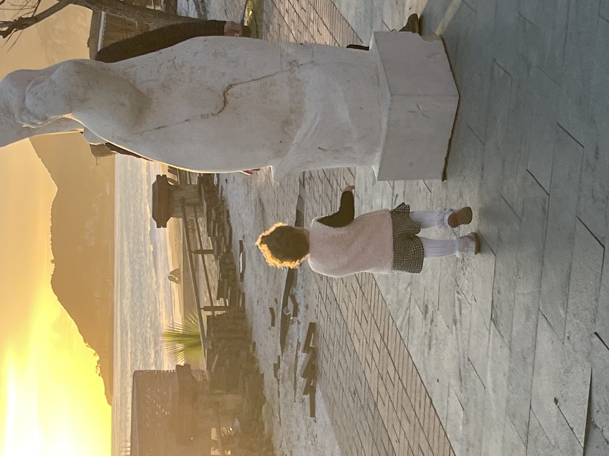 A little girl stands on a stone-paved path beside a white sculpture, likely in Alanya, Turkey, given the coastal view and mountainous terrain visible in the background. The scene is bathed in the golden light of either sunrise or sunset. The child is wearing white tights, brown shoes, and a dress with a pink sweater or vest. The sculpture is large and possibly made of marble or a similar material. The background includes a body of water, a beach, buildings, and a mountain silhouetted against the bright sky. There's a wooden fence or barrier along the path.