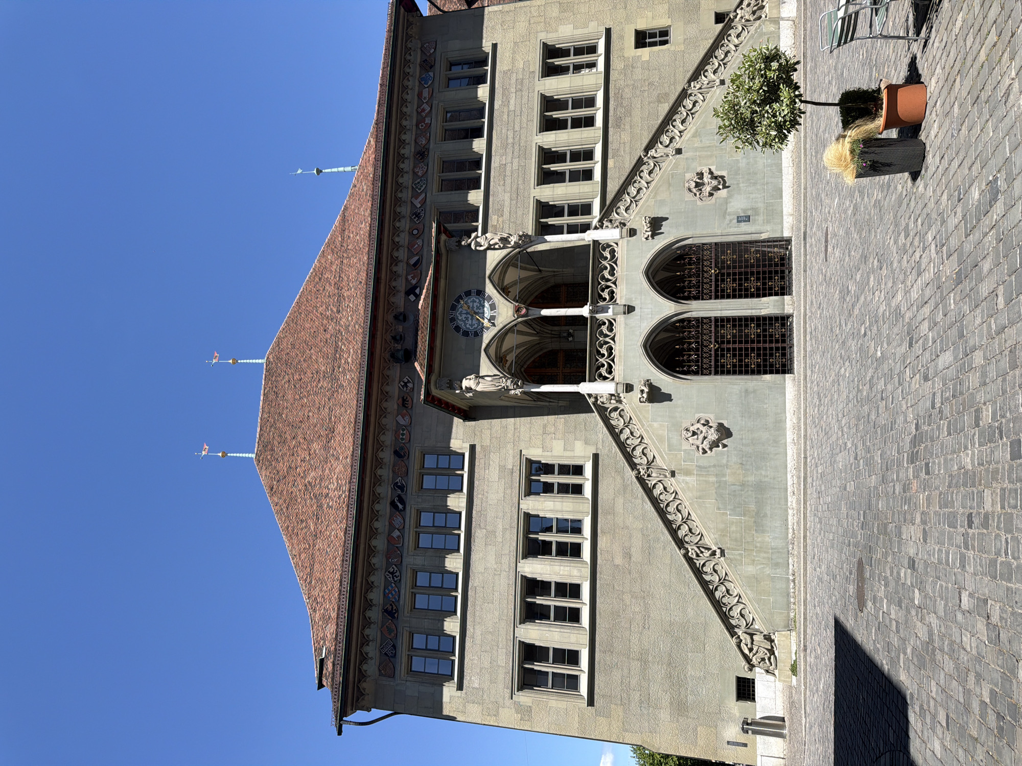 The image shows the Zytglogge, a clocktower and former medieval guard tower in Bern, Switzerland. The clocktower is a significant landmark in the city. The tower is made of stone and has a red tiled roof with several spires. A large clock face is visible on the front of the tower. There is a set of stairs leading up to the entrance of the tower, with ornate railings. The ground in front of the tower is paved with cobblestones. There are two potted plants and a table with chairs in the foreground. The sky is clear and blue.