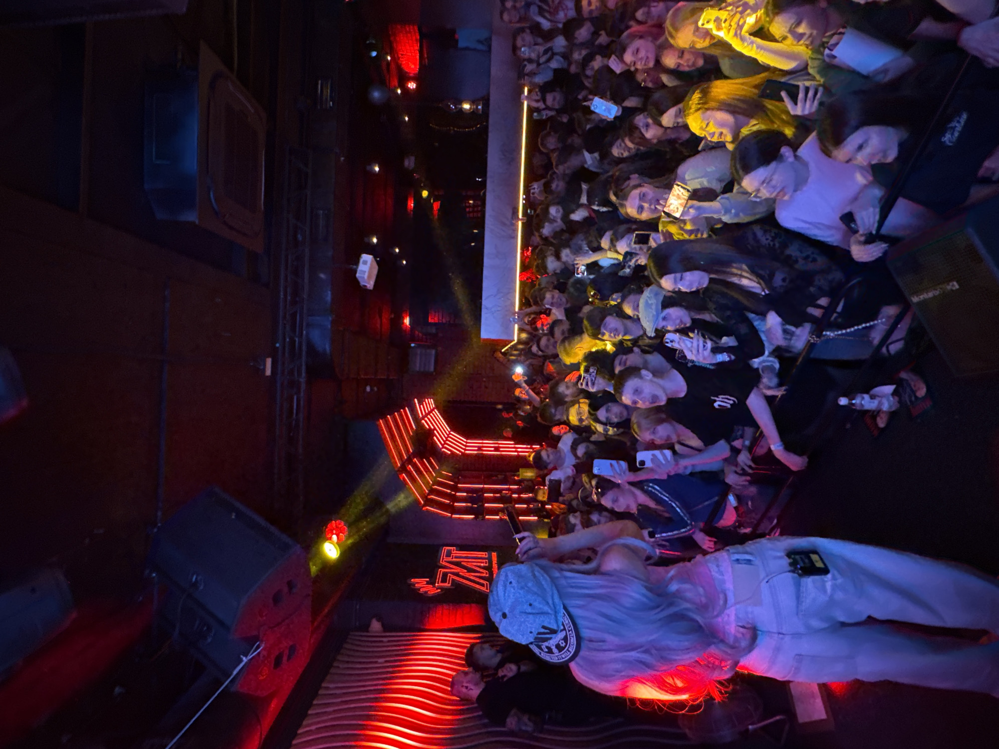 The image depicts a lively concert or live performance taking place at night in a dark venue in Khabarovsk, Russia. The scene is captured from an elevated perspective, looking down onto the stage and the audience.
In the foreground, a performer is visible from behind, facing the crowd. They have long, light-colored hair, wear a light-colored baseball cap backward, a light-colored top, and light pants, with a small electronic device clipped to their back pocket. The performer is holding a microphone, actively engaging with the audience.
The stage area is brightly lit with dramatic red and yellow spotlights, casting strong illumination and shadows. Red strip lights highlight the steps leading up to the stage. On the wall behind the performer, a neon-red logo or stylized text, resembling "M7" or "W7," is prominently displayed. Speakers are positioned on the stage, with text like "MONITOR 10 C" possibly visible on one. Another person is partially visible lying on the stage floor near the performer.
A large crowd of people, mostly young adults, fills the area beyond the stage, packed closely together and facing the performer. Many audience members are holding up smartphones, their screens casting a glow on their faces as they record or photograph the event. Their expressions appear engaged and excited, contributing to the energetic atmosphere of the concert. The venue's walls are dark, with minimal ambient lighting, emphasizing the illuminated stage and the glowing phone screens of the crowd.