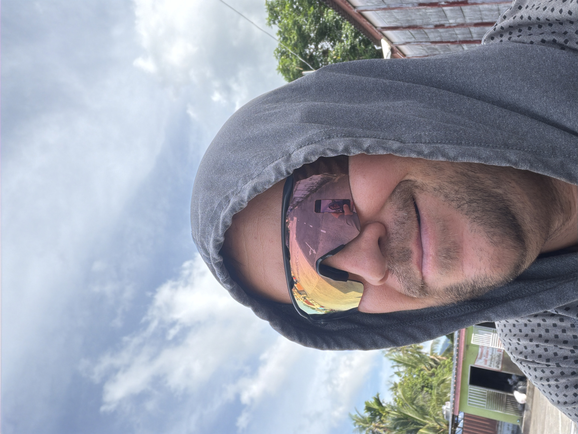 A man is wearing a gray hooded sweatshirt and sunglasses, with the sky and clouds visible in the background. The sunglasses reflect a street scene with buildings and trees. He appears to be outdoors in a tropical or subtropical environment, possibly in Baan Mae Nam, Thailand, given the presence of palm trees. The overall impression is a casual, candid moment.