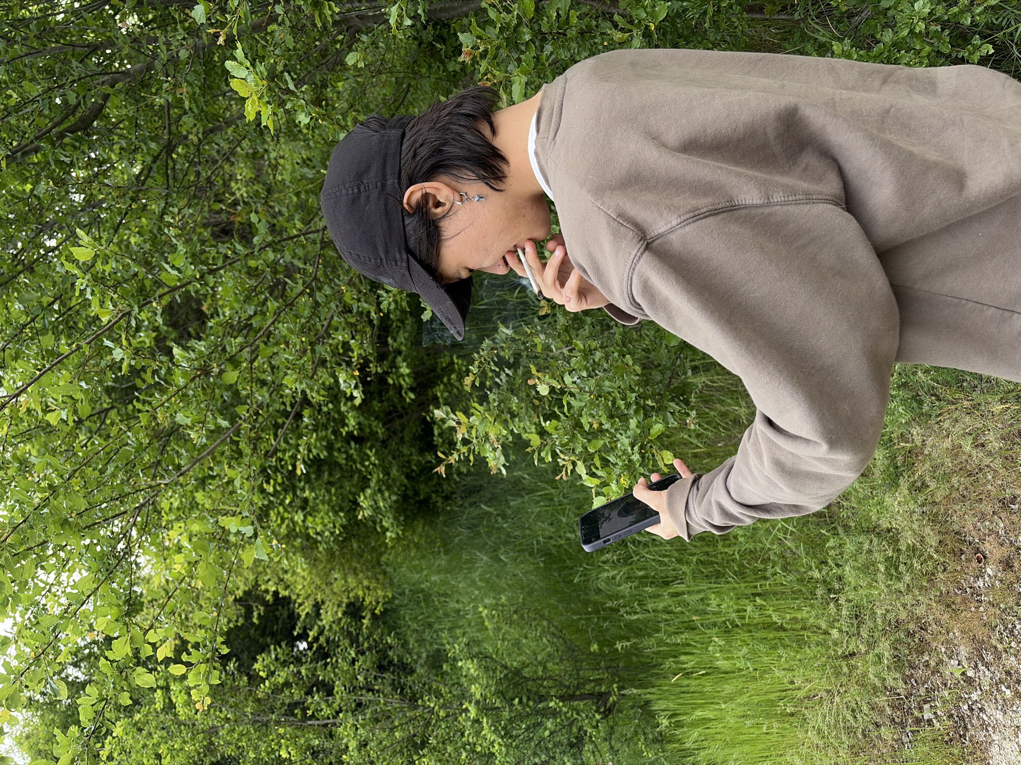 A young person stands outdoors on a grassy path bordered by trees and vegetation. The person wears a backwards black baseball cap and a tan sweatshirt, and holds a cell phone in their right hand. They are smoking a cigarette. The location is unspecified, and there is no evidence to indicate it is in the city of Sofia, Bulgaria.