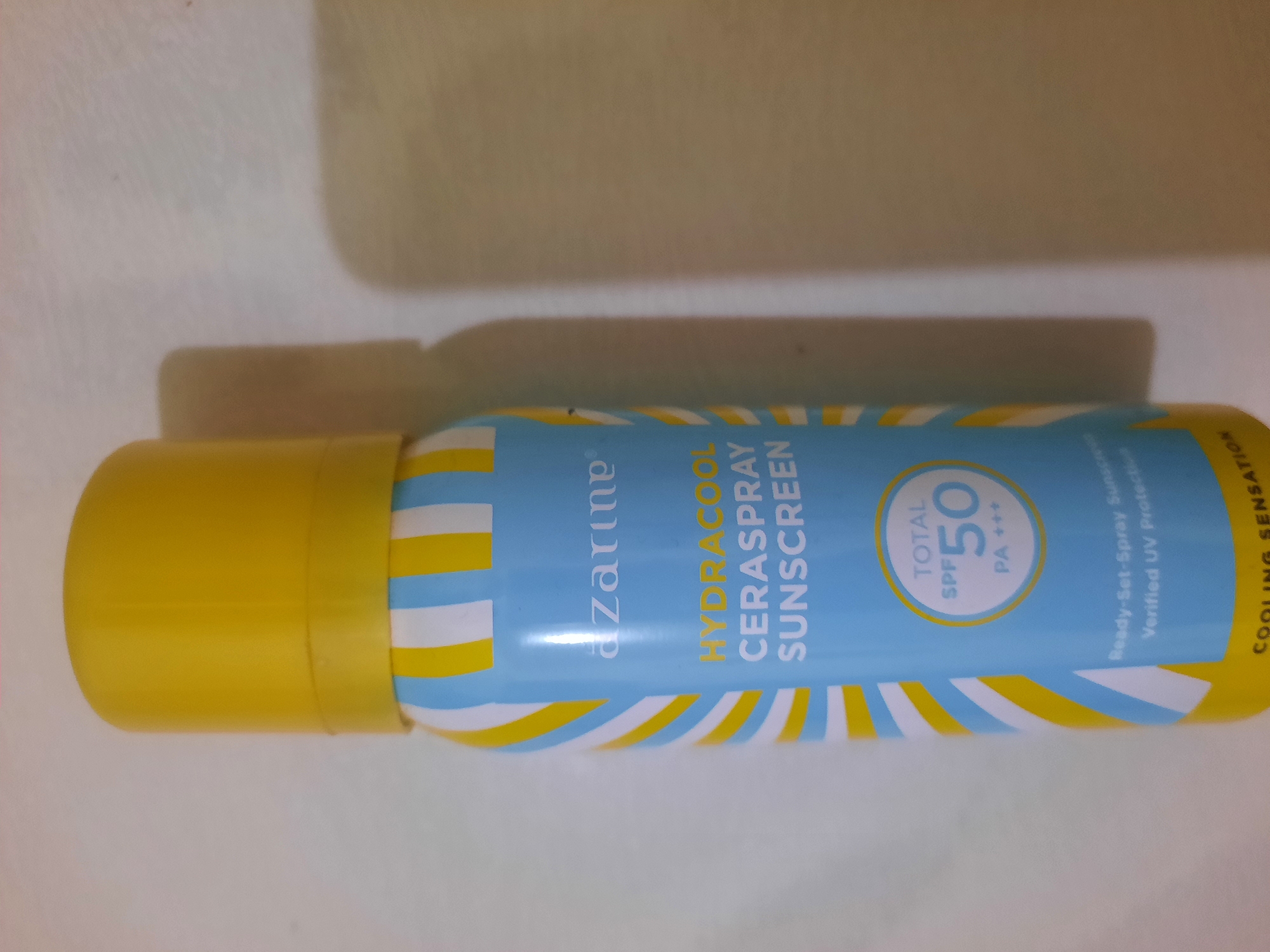 An overhead shot shows a sunscreen spray can. The can is light blue with diagonal yellow and white stripes. The text on the can reads "AZARINE HYDRACOOL CERASPRAY SUNSCREEN" and "TOTAL SPF 50 PA+++". The can has a yellow cap. The background is a light-colored, textured surface that appears to be a table or countertop. The lighting casts a soft shadow from the can onto the surface. The image was taken in Lhokseumawe, Indonesia.