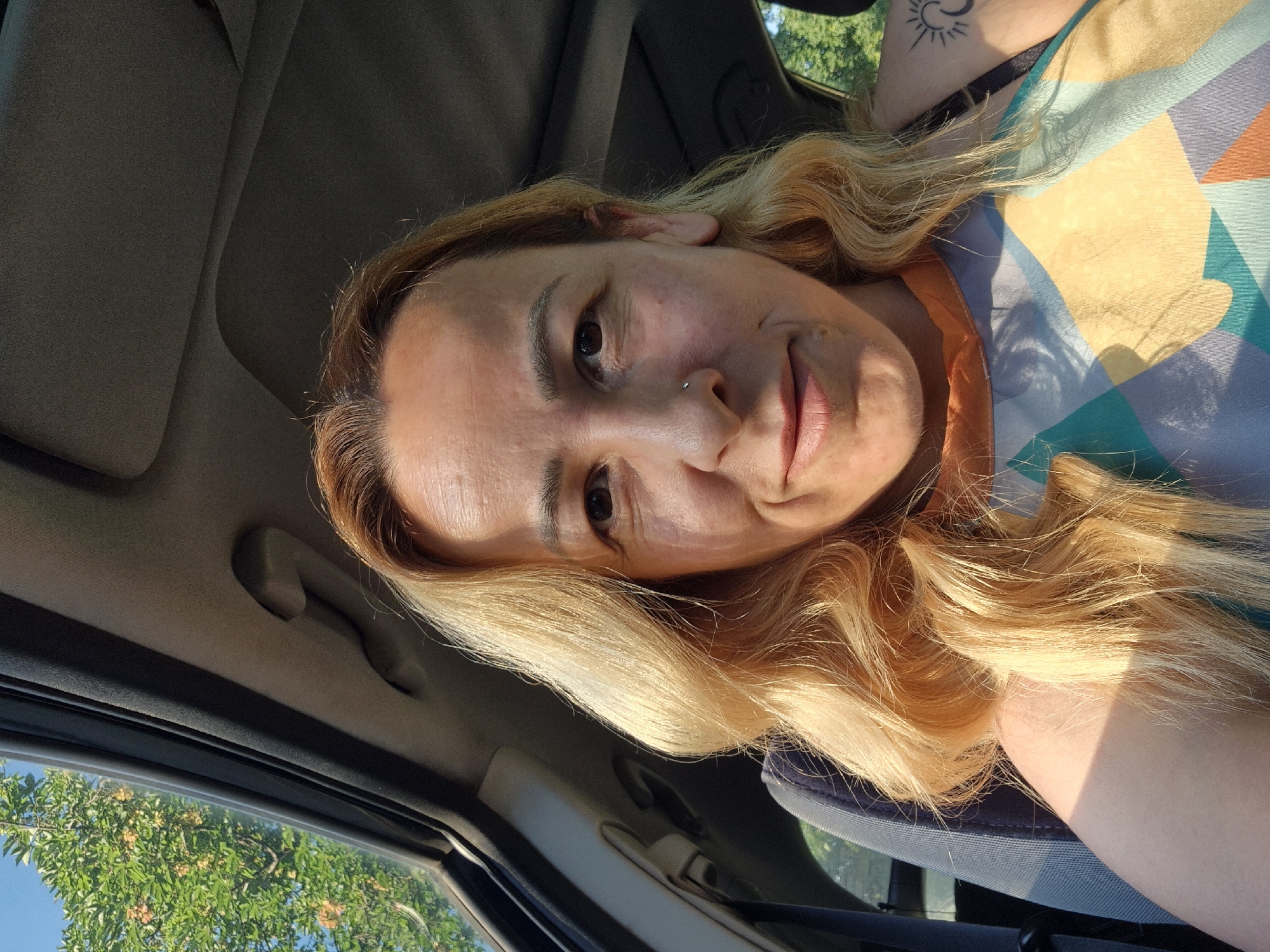 A woman is taking a selfie in a car, with her face and upper body visible. She has blonde hair and is smiling. The car's sunroof is open, and trees and a blue sky are visible in the background. She is wearing a colorful patterned shirt.