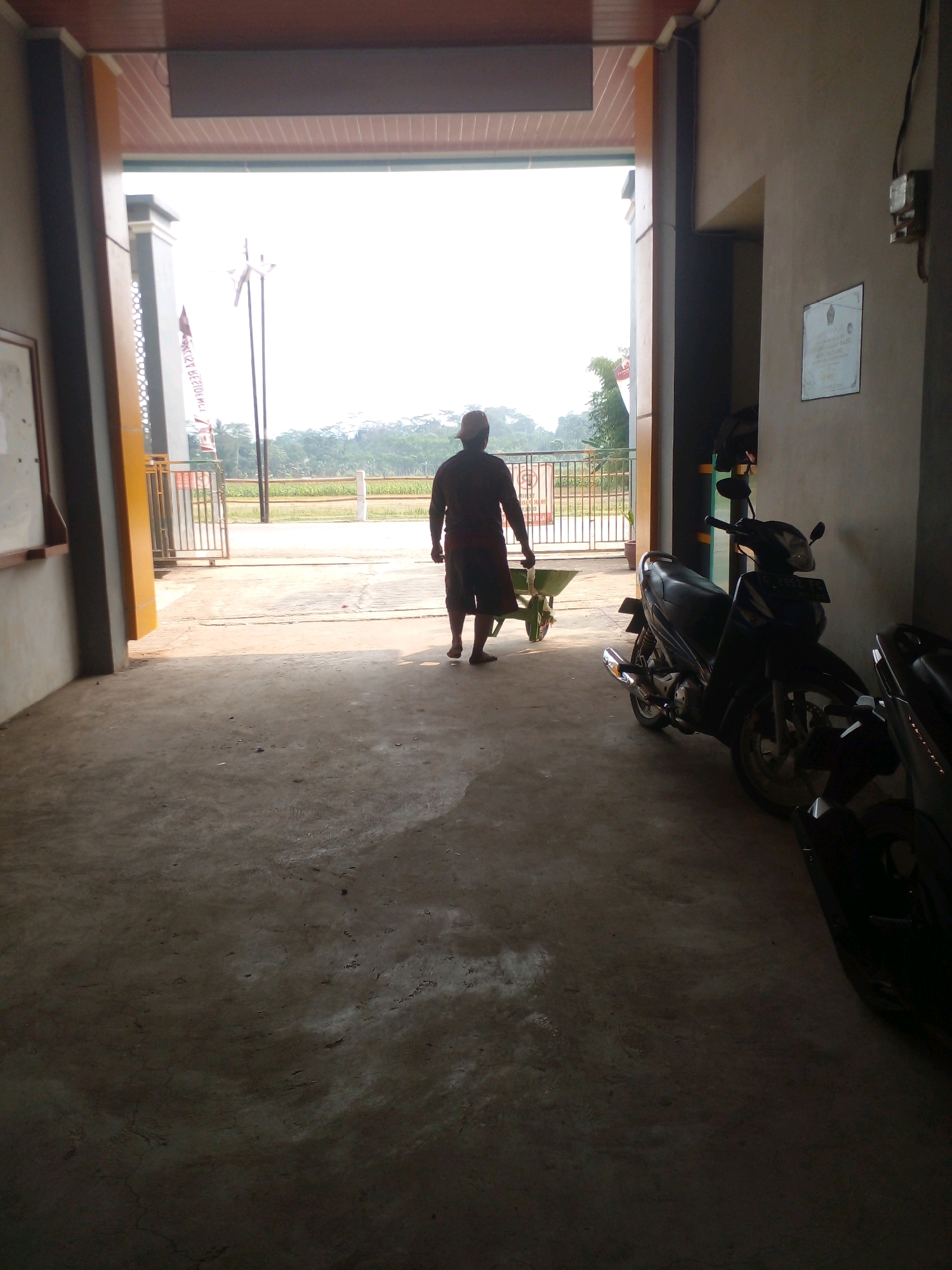 The image depicts a man from behind, wearing a dark t-shirt and shorts, pushing a green wheelbarrow towards an open doorway. He is walking on a concrete surface. To the right, two motorcycles are parked. The background shows a bright, outdoor scene with a fence, vegetation, and hills under a clear sky. The setting appears to be an outdoor area associated with a building, possibly in a rural or semi-urban location in Banyuputih, Indonesia, given the presence of motorcycles and the general landscape.