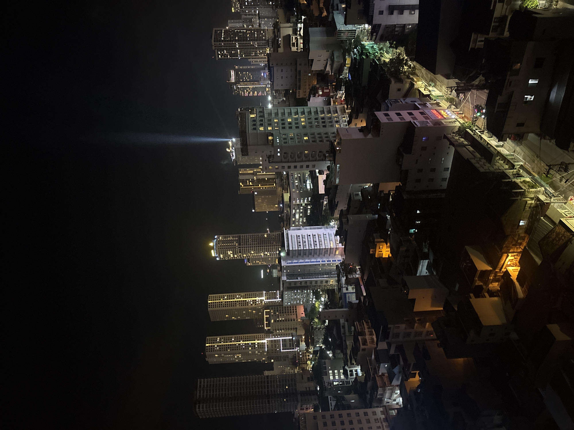 The image depicts a nighttime urban scene in Nha Trang Ward, Vietnam. High-rise buildings dominate the cityscape, illuminated with numerous lights. A bright beam of light emanates from one of the structures. Lower-rise buildings and structures are interspersed among the skyscrapers. The environment is dark, save for the artificial lights illuminating the buildings and streets. There are no people visible in the image.