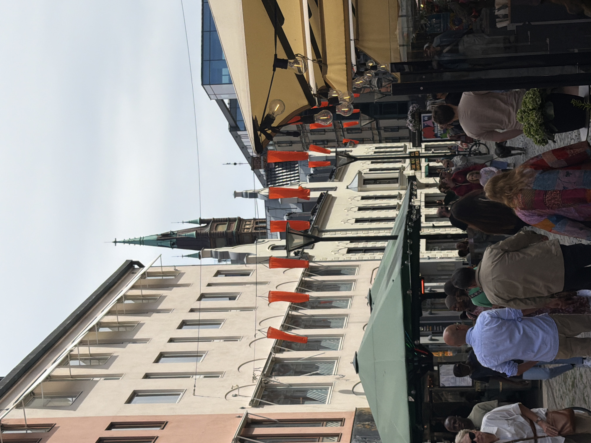 The image depicts a street scene in Malmö, Sweden, likely during daytime given the lighting. The street is lined with buildings of varying architectural styles, including older structures with light-colored facades and a modern building with a glass facade. A prominent church spire with green detailing is visible in the background.
The street is moderately crowded with people walking in various directions. Some individuals are dressed casually, while others appear to be patrons of outdoor seating areas. Several people are walking with their backs to the camera, making their features indistinguishable. Visible elements include outdoor umbrellas or awnings, string lights, and decorative orange banners hanging overhead. The ground appears to be paved with cobblestones or similar material. In the foreground, there are outdoor seating structures, possibly for a restaurant or cafe, with umbrellas providing shade.
The overall impression is that of a bustling urban environment, possibly a commercial or pedestrian zone, with a mix of historical and contemporary elements.
