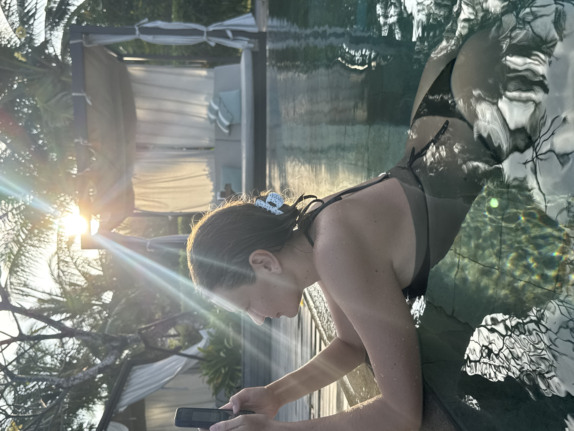 A woman in a black swimsuit is leaning on the edge of a swimming pool and looking at a smartphone in Kuta Selatan, Indonesia. The sun is low, causing strong light flares through the trees. There's a white canopy bed near the pool. The water reflects the trees above.