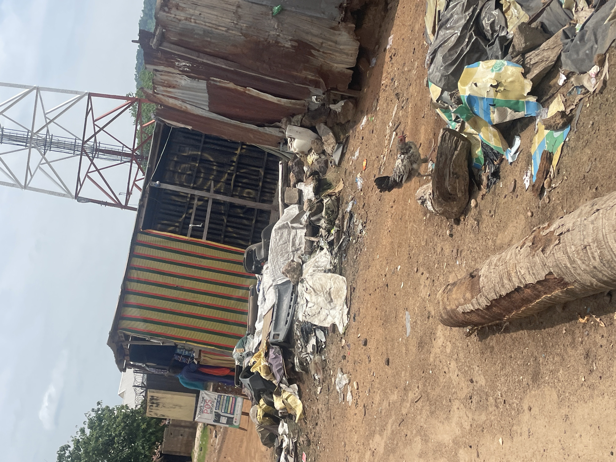 In Lokoja, Nigeria, a scene of disarray unfolds near a telecommunications tower. A makeshift shop, constructed from corrugated metal and fabric, leans precariously. The shop displays colorful vertical stripes of green, red, and yellow. A sign in front of the shop reads "GOD'S TIME ENTERPRISE, POS SERVICE AVAILABLE." Two individuals are visible near the entrance, one wearing a blue garment and the other a reddish-orange outfit.
In front of the shop, a significant amount of debris is scattered across the reddish-brown dirt ground. This debris includes what appears to be car parts, white and yellow sheeting, plastic bags, and other miscellaneous refuse. Further into the scene, a chicken with speckled grey and white plumage stands on a weathered piece of wood. To the right, more piles of black plastic sheeting and torn colorful material contribute to the overall cluttered appearance. A fallen, weathered log lies in the foreground, adding to the rustic and somewhat neglected environment. The telecommunications tower, a prominent structure of red and white metal lattice, looms in the background, its presence a stark contrast to the ground-level disarray. The sky is overcast with scattered clouds.