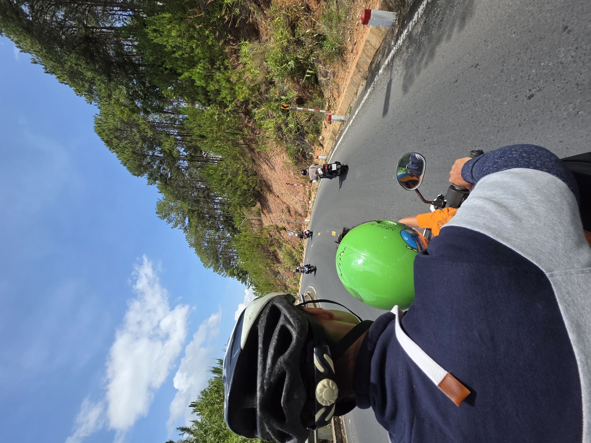 The image shows a first-person perspective while riding a scooter on a road in Thôn 5, Vietnam. In the foreground, two people wearing helmets are visible. The person closest to the camera is wearing a dark gray bicycle helmet, and the second person wears a bright green helmet. The person filming is wearing a blue and gray hoodie. Beyond them is the gray road with other scooters farther ahead. A grassy, wooded hillside borders the right side of the road, with trees and a clear, blue sky with clouds on the left. Roadside barriers are visible intermittently on the roadside.