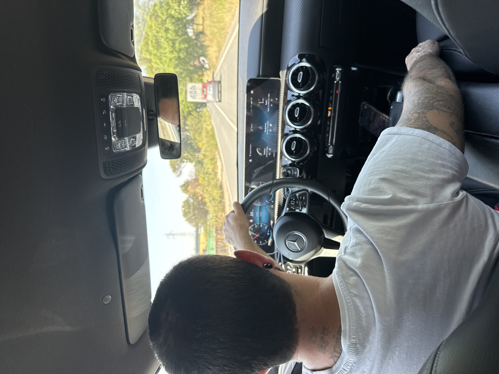 The image depicts the interior of a car, specifically the driver's seat area, with a person driving. The person has short, dark hair, wearing a white shirt and tattoos on his left arm. His hands are on the steering wheel, and the dashboard features a digital display with navigation information. The car is traveling on a road, and scenery is visible through the windshield including trees, a road, a truck and buildings. The scene suggests that the car is likely located in a suburban or rural area. Based on information from the question, this is in Ardea, Italy. The interior of the car shows a modern design with climate control vents and other dashboard features. The rearview mirror is visible in the upper center of the image.