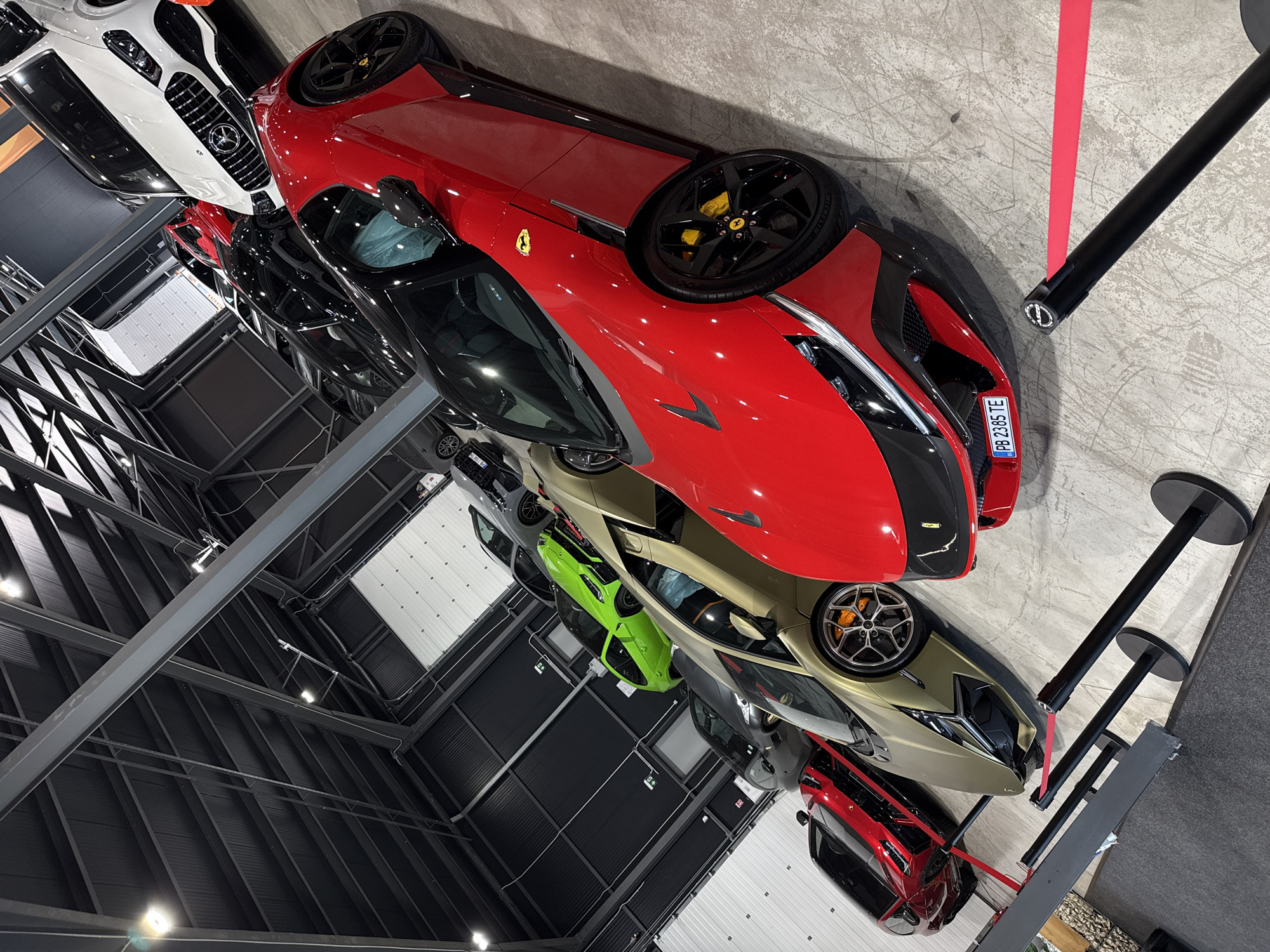The image shows a collection of luxury cars, primarily sports cars, parked indoors. The cars are arranged in rows, with a prominent red Ferrari in the foreground. Other visible cars include a gold Lamborghini, a bright green hatchback, and several darker colored sedans and sports cars. In the background, a white Mercedes-Benz SUV is also visible. The environment appears to be a showroom or a garage, characterized by a high ceiling with exposed industrial-style beams and lighting fixtures. The floor is concrete, and there are stanchions with red velvet ropes, suggesting a display or exhibition setup. The overall impression is one of a high-end automotive exhibition. There are no people visible in the image. The license plate on the red Ferrari reads "PB 2385 TE". The location is specified as Plovdiv, Bulgaria.