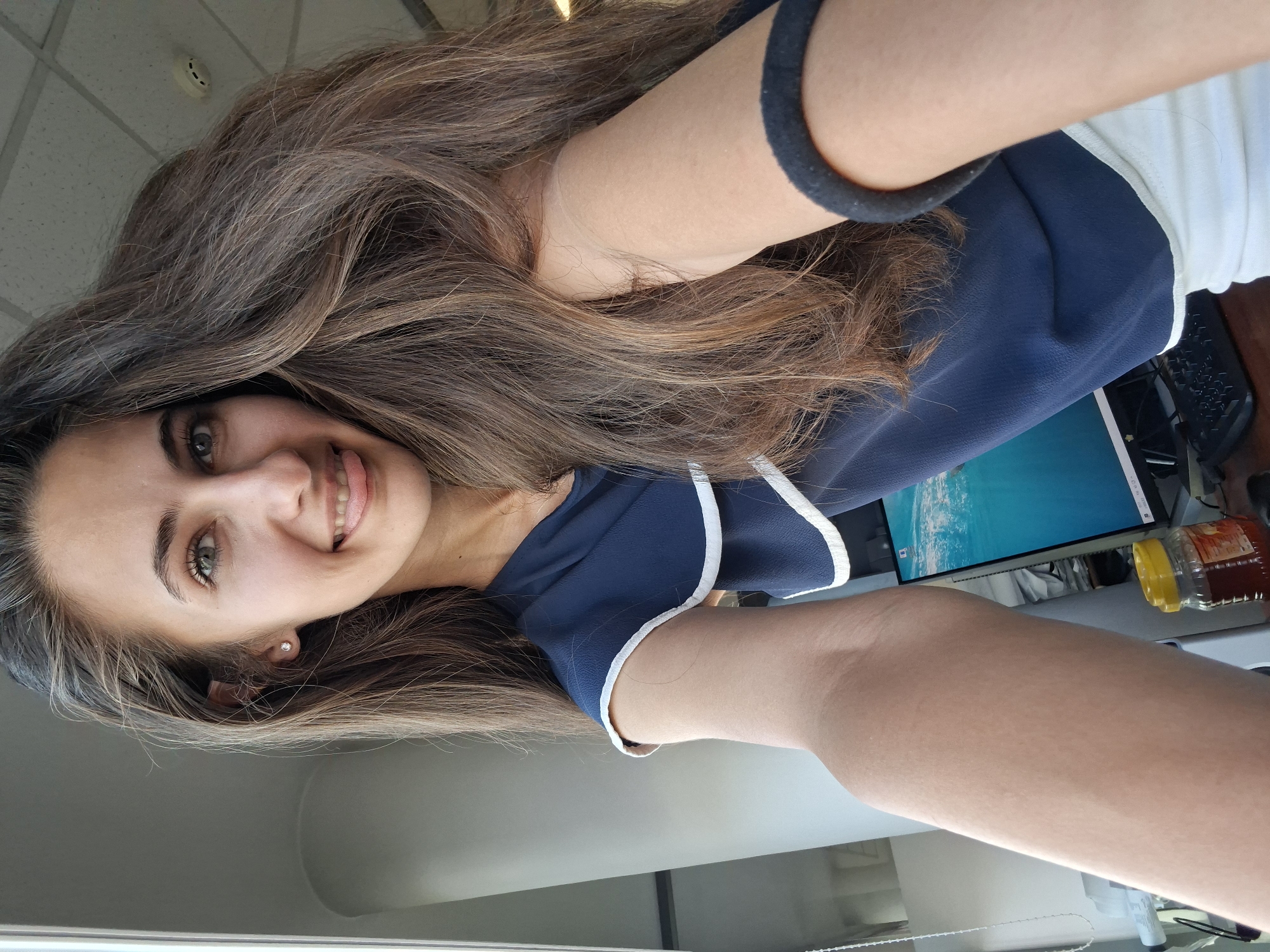 In an indoor setting, a young woman with long, brown hair is taking a selfie. She is wearing a dark blue top with white trim and white shorts. Her right arm is extended forward, holding the camera. She is smiling and sticking her tongue out slightly. Behind her, a computer monitor displays a bright blue image, and a jar of honey sits on a desk. The scene appears to be a home office or a workspace.