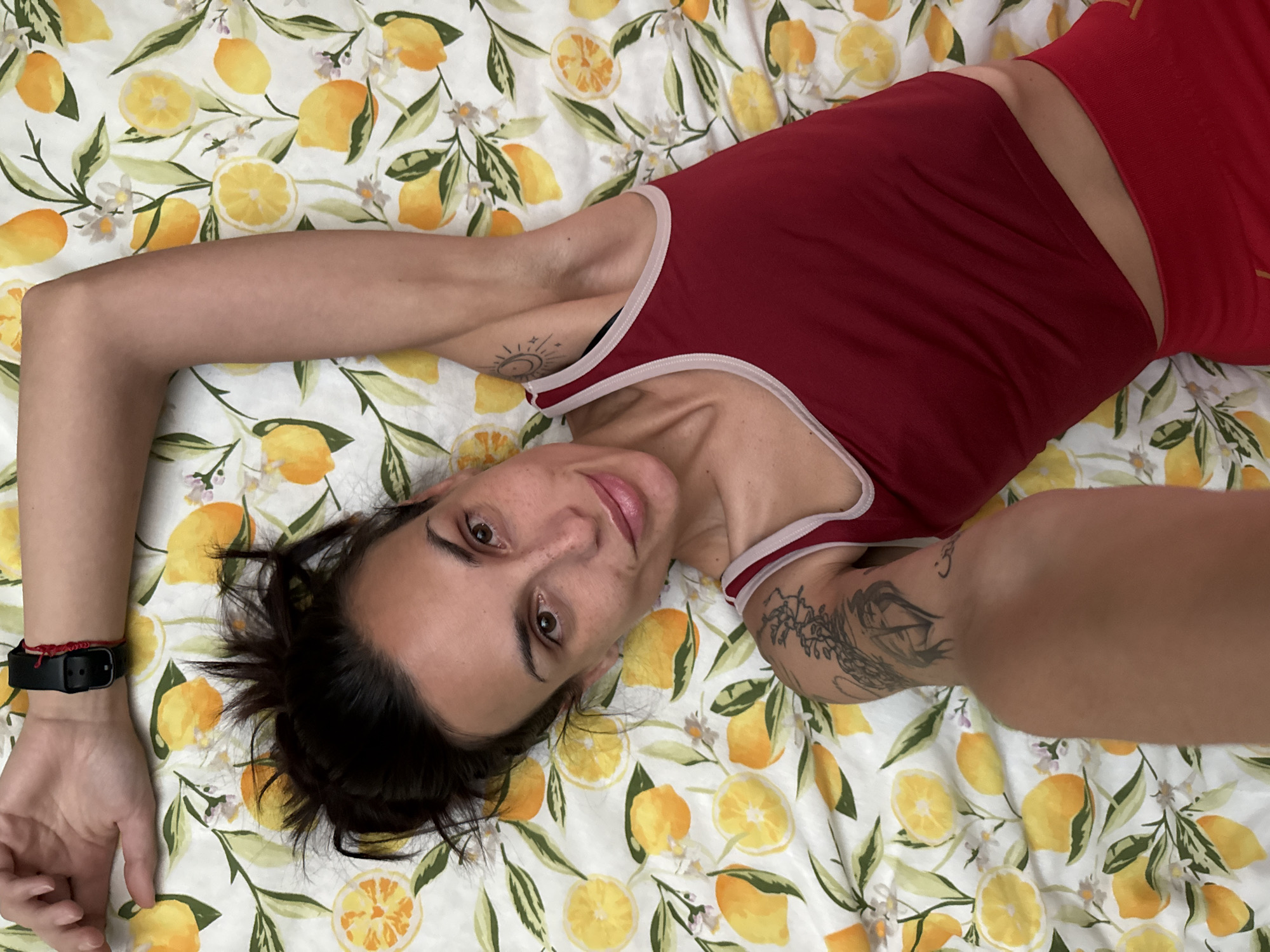 A person is lying on a bed with a lemon-patterned sheet. The person is wearing a red tank top with white trim and red shorts. The person has dark hair pulled back and is looking at the camera with a slight smile. The person has tattoos on their arms. The person is wearing a black watch on their left wrist and a red bracelet on their right wrist. The location is likely indoors, but the specific city of Pleven, Bulgaria, cannot be determined from the image alone.