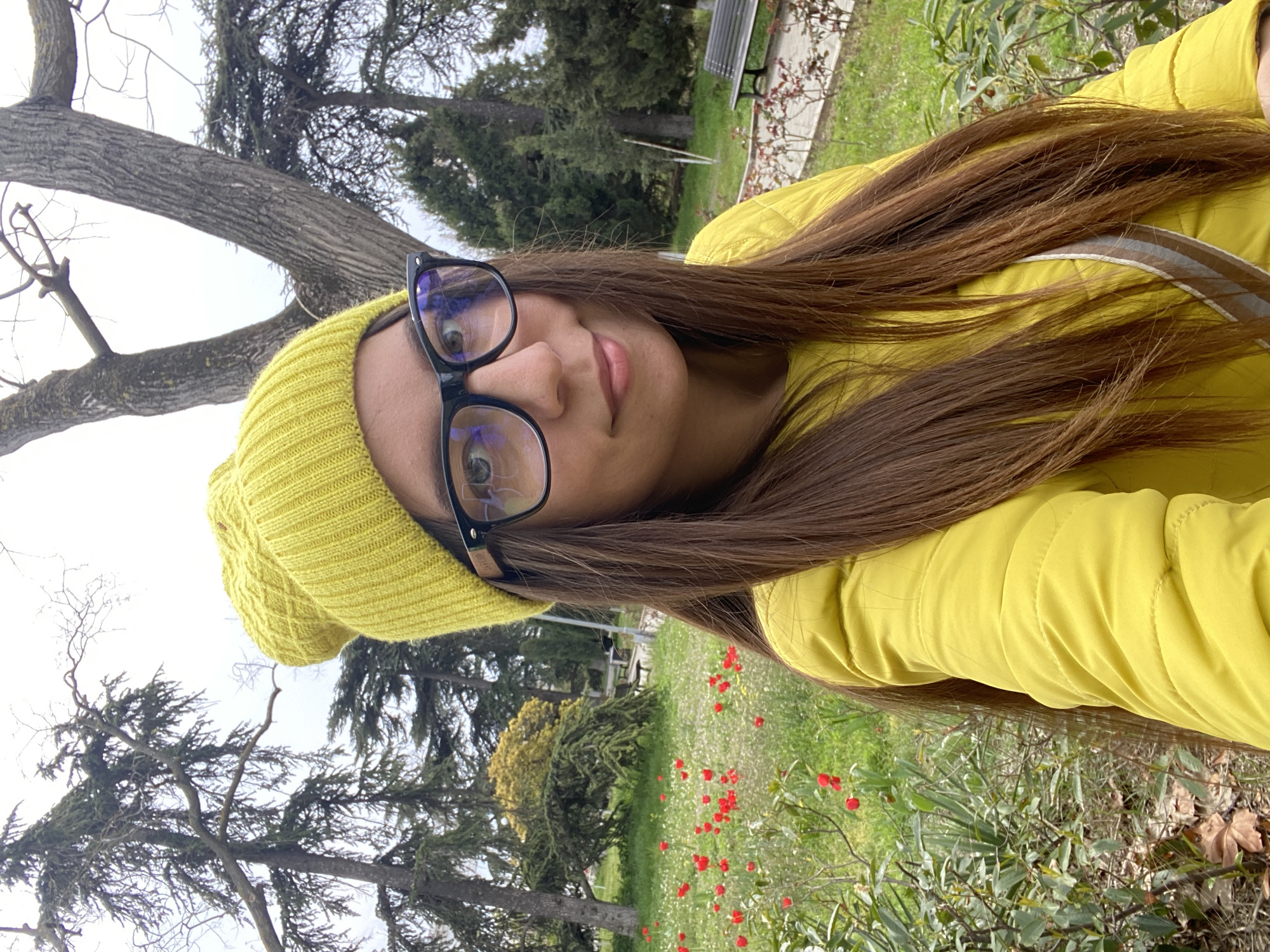A young woman wearing a yellow beanie and glasses is taking a selfie outdoors in a park-like setting. She is also wearing a yellow jacket. The environment includes trees, grass, and red flowers. In the background, a bench can be seen, suggesting a public park or garden location. While I am unable to confirm that this picture was taken in Sevastopol, Ukraine, I can say that this image features an outdoor setting that resembles natural landscapes that can be found there.