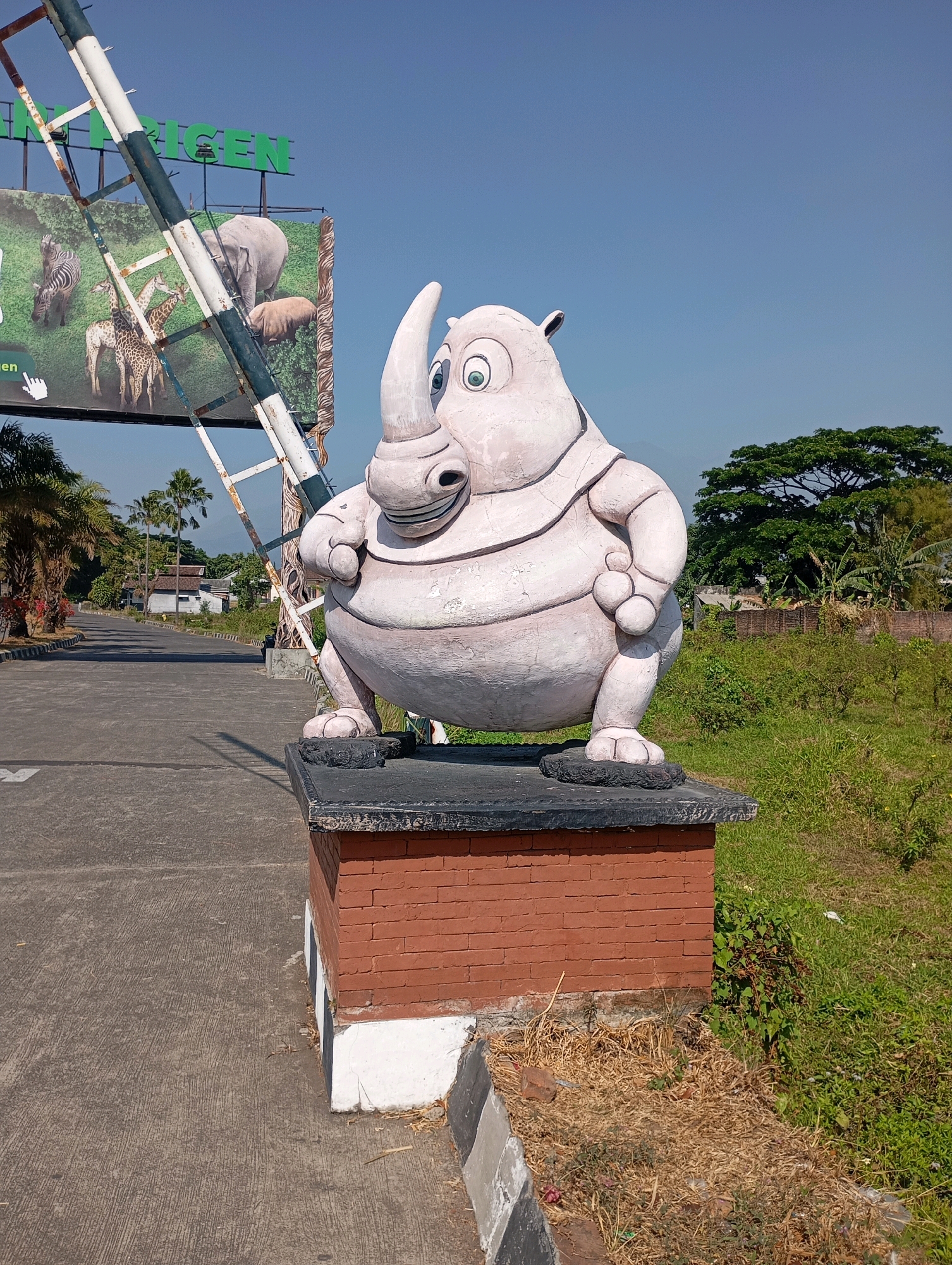 A large, cartoonish rhino statue stands on a brick pedestal by a roadside in Lemahbang, Indonesia. The statue is light pink with large, round eyes, a prominent horn, and a plump body. It is positioned on the right side of the image, facing left. To the left of the statue, a large billboard displays images of wildlife, including zebras, giraffes, and an elephant, and the text "Lemahbang" is visible in green lettering. A metal structure, possibly part of an entrance gate, leans across the billboard. The background shows a road leading into the distance, lined with palm trees and residential buildings. Green vegetation and a brick wall are visible to the right of the road. The sky is clear and blue.