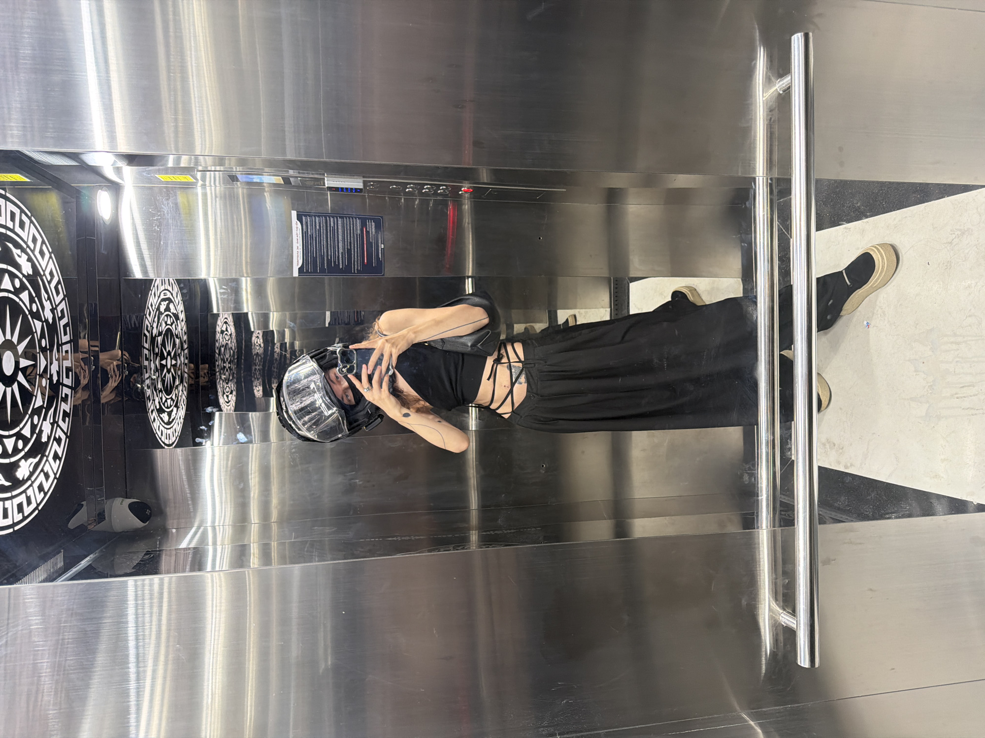A woman wearing a clear helmet and dark clothing is standing in an elevator taking a photo with her phone. The elevator has stainless steel walls and a decorative panel with geometric patterns. The floor is black and white marble. The location is likely in the city of Đà Nẵng, Vietnam, based on the geometric patterns in the elevator which resembles elements of Vietnamese art.