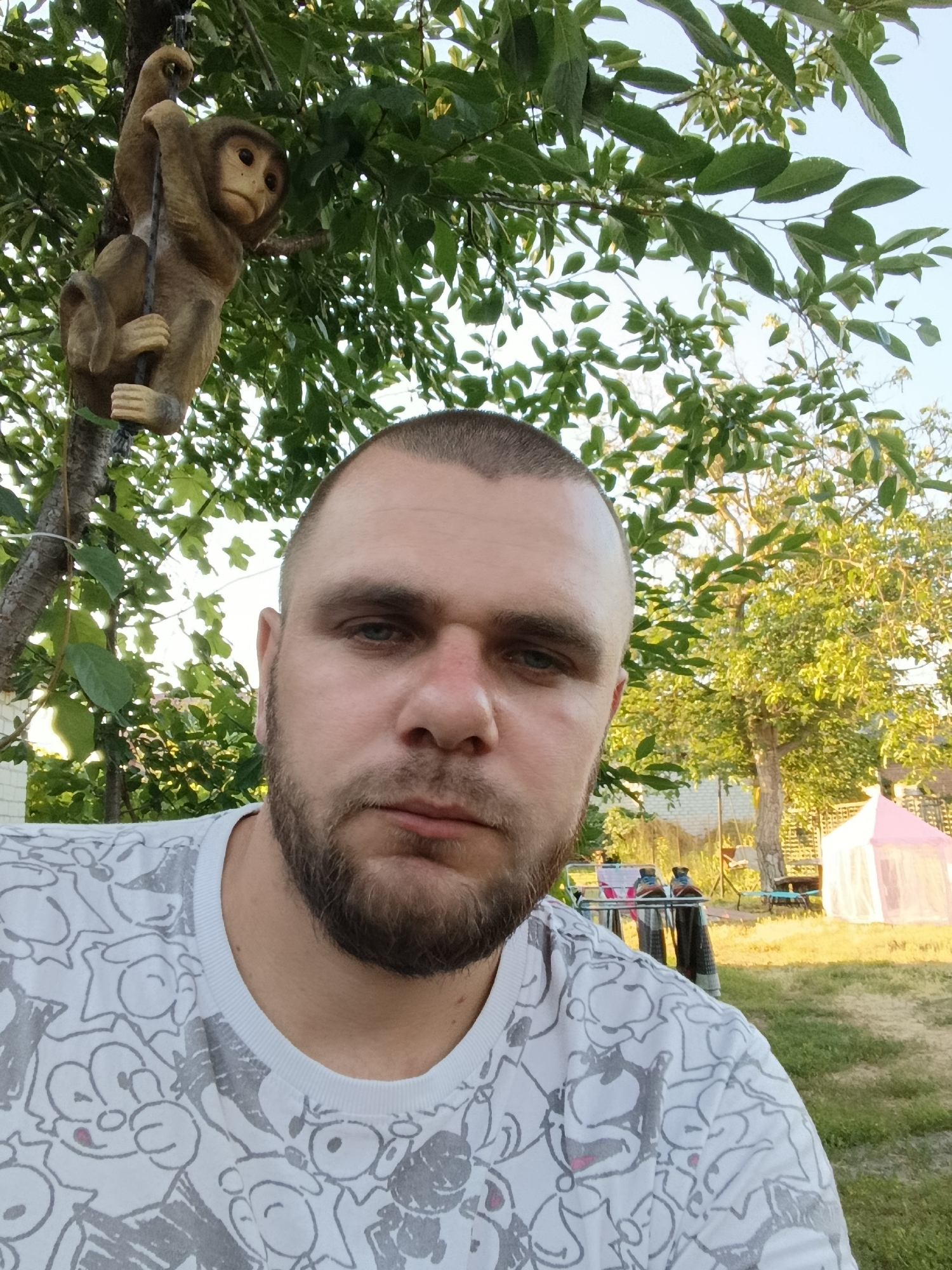 In an outdoor setting, a man with a shaved head and a beard takes a selfie. He is wearing a white t-shirt with a cartoonish gray pattern. To his left, a decorative monkey figure hangs from a tree branch by its tail, suspended by a dark string. The background reveals a backyard with a clothesline, a pink tent, and lush green trees. The lighting suggests it is late afternoon or early evening.
