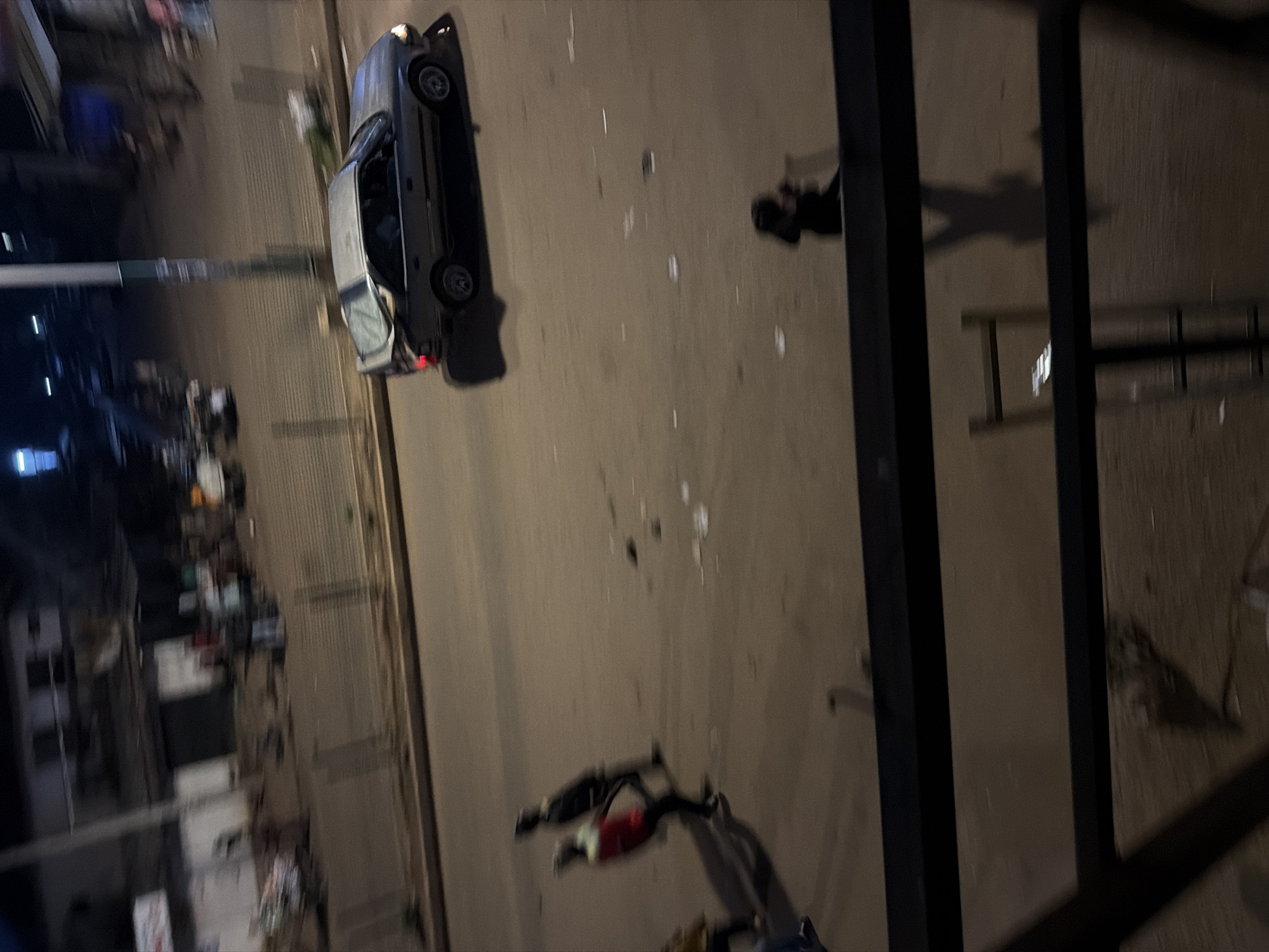 An aerial, slightly blurred, night-time photograph shows a street scene in Bukuru, Nigeria. A silver sedan is driving on the road. Two individuals, appearing to be young men, are walking on the sidewalk. One is wearing a red shirt and black pants, and the other is in a dark outfit. A third individual, partially visible, is also walking on the sidewalk. There are scattered pieces of debris on the ground, including what appears to be litter. The environment includes buildings in the background and a metal fence separating the sidewalk from the road. The overall lighting suggests it is dusk or night.